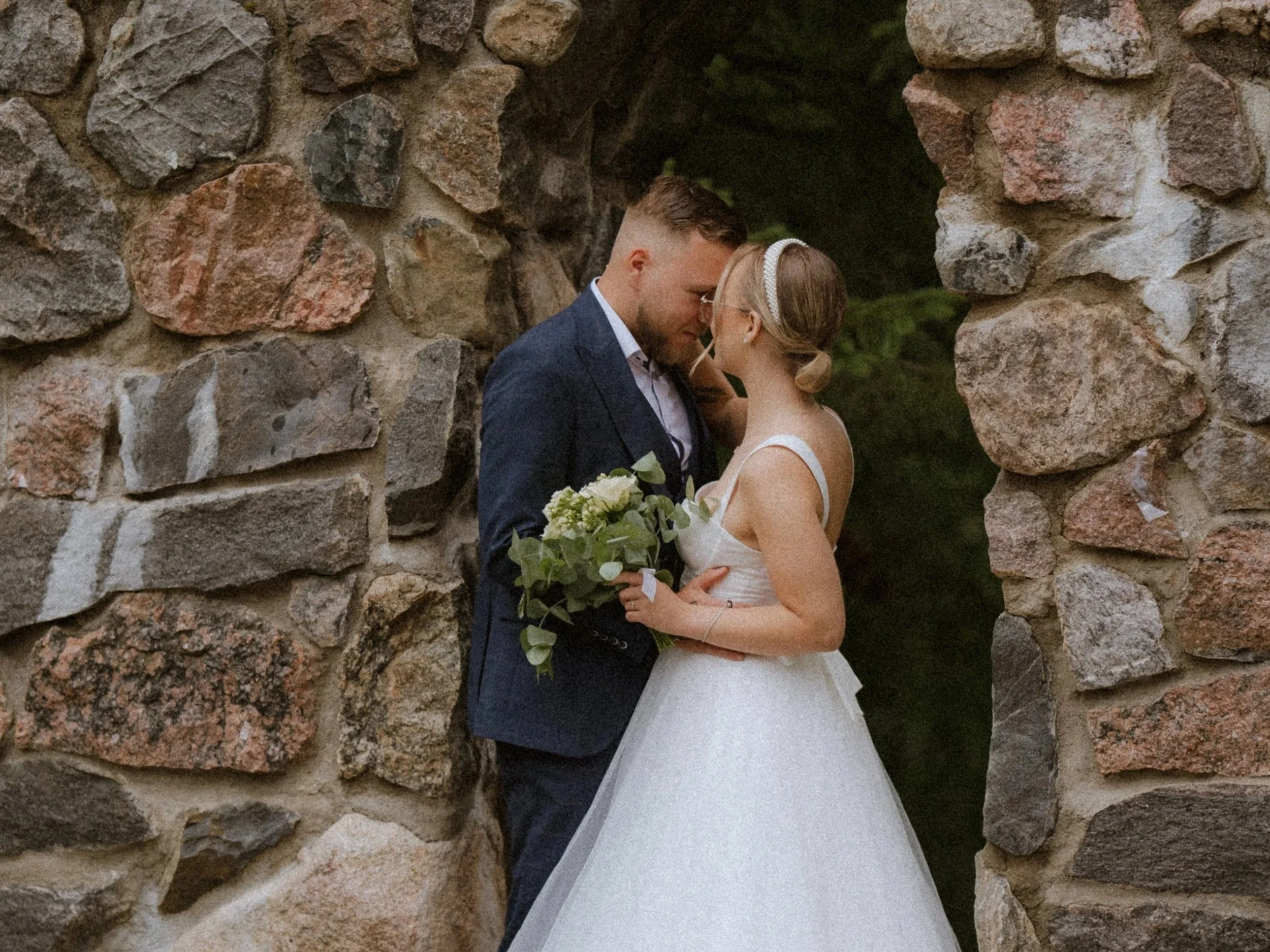 A bride and groom standing close, with foreheads touching, inside a stone archway. The bride is holding a bouquet of flowers and wearing a white wedding dress with a headband. The groom is dressed in a navy blue suit with a white shirt.