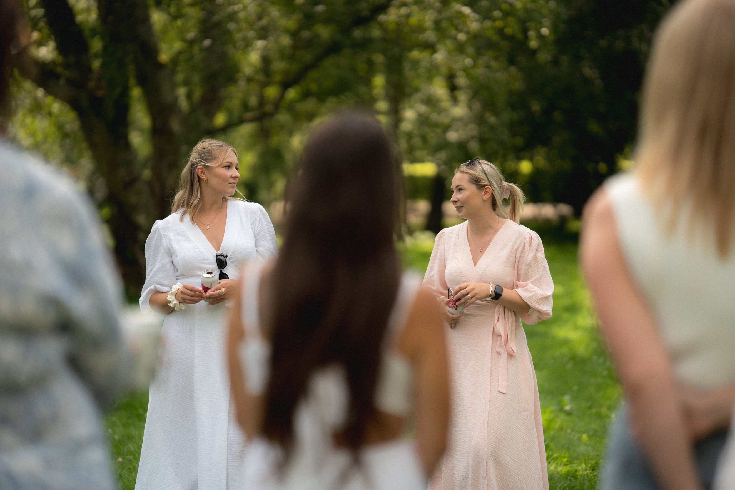 Two women in light-colored dresses engaged in conversation outdoors among other people in a park setting with green trees.