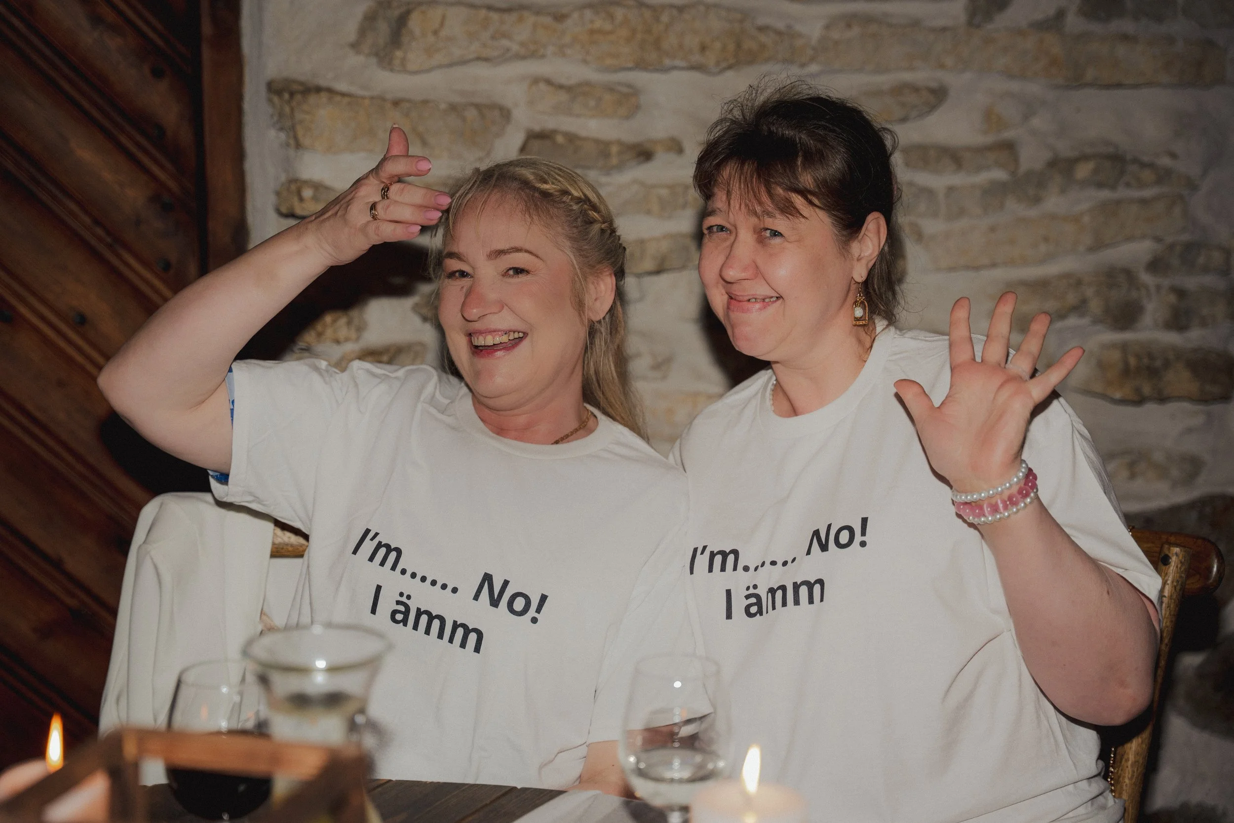 Two women sitting at a table, smiling and waving, wearing matching t-shirts with humorous text, in front of a rustic brick wall.