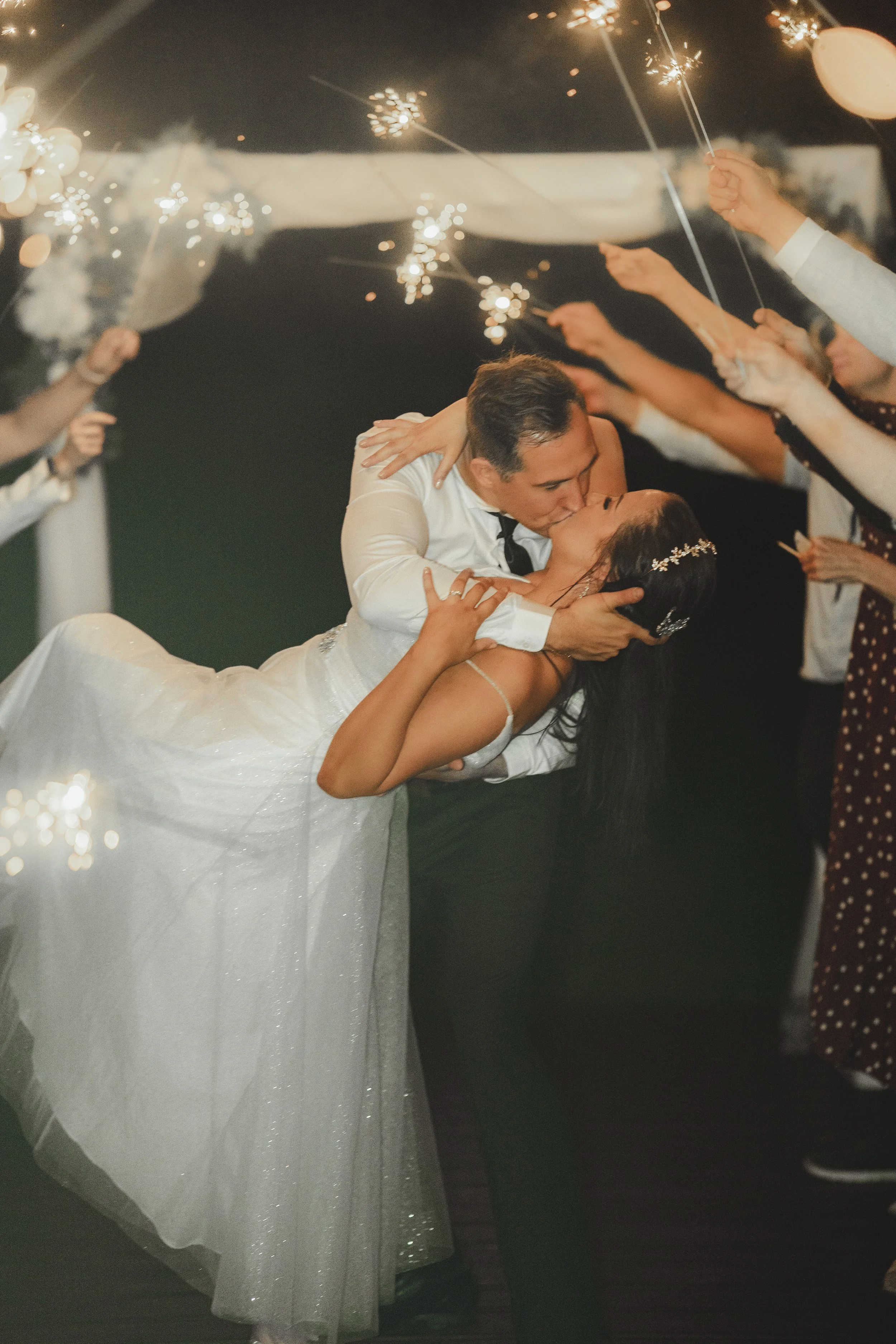 A bride and groom kiss as friends and family hold sparklers overhead at a wedding celebration.