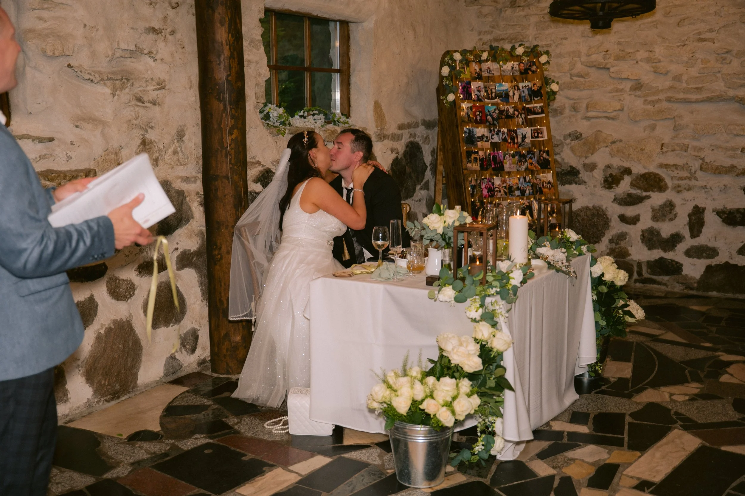 A bride and groom share a kiss at their wedding reception, seated at a decorated table with flowers, candles, and photo displays in a rustic stone-walled room.