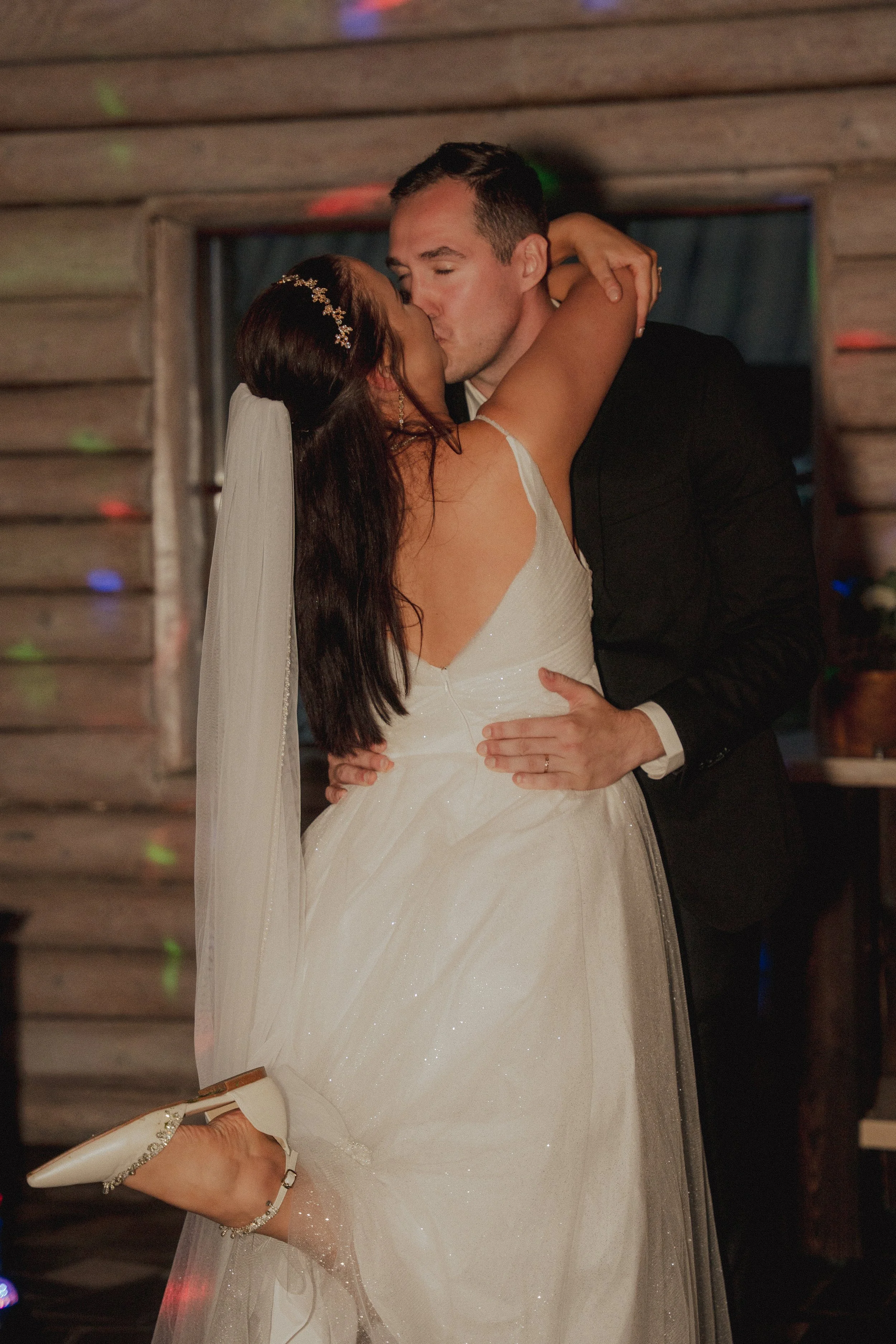 A bride and groom share a kiss during their wedding reception, with the bride raising one leg and holding the groom tightly, in a rustic wooden venue.