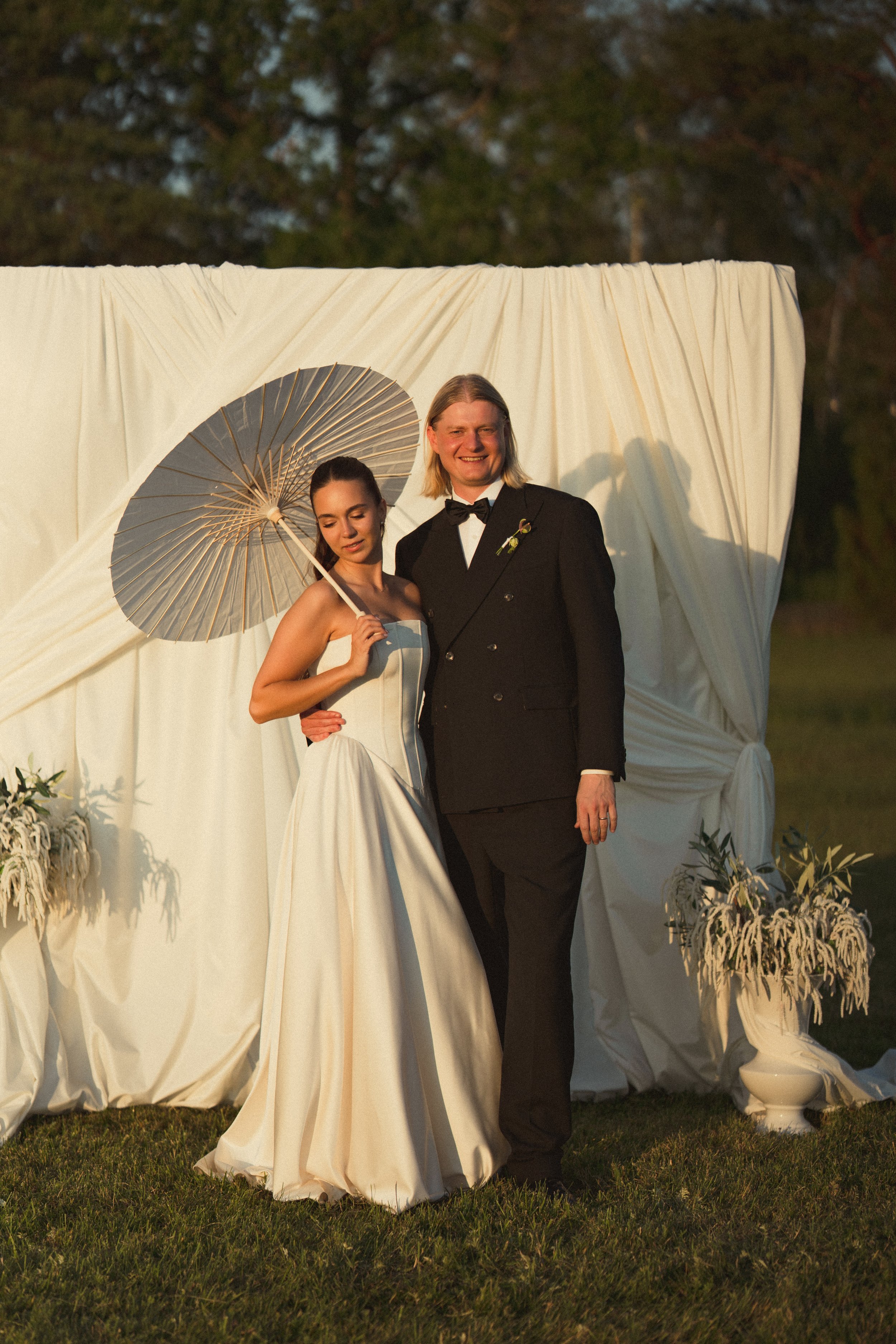 A bride and groom stand close together outdoors at sunset, smiling, with a white fabric backdrop and floral arrangements nearby. The bride is in a strapless white wedding gown holding a silver parasol, and the groom wears a black tuxedo with a bow ti