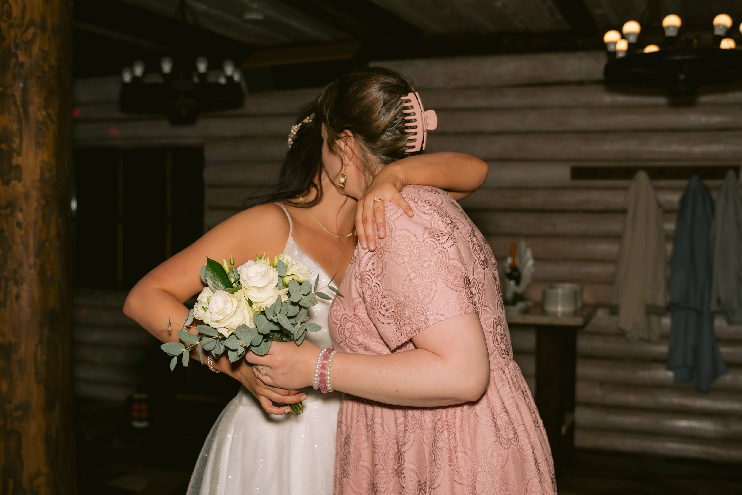 A bride hugging a woman in a pink dress inside a rustic wooden venue, with the bride holding a bouquet of white roses and greenery.