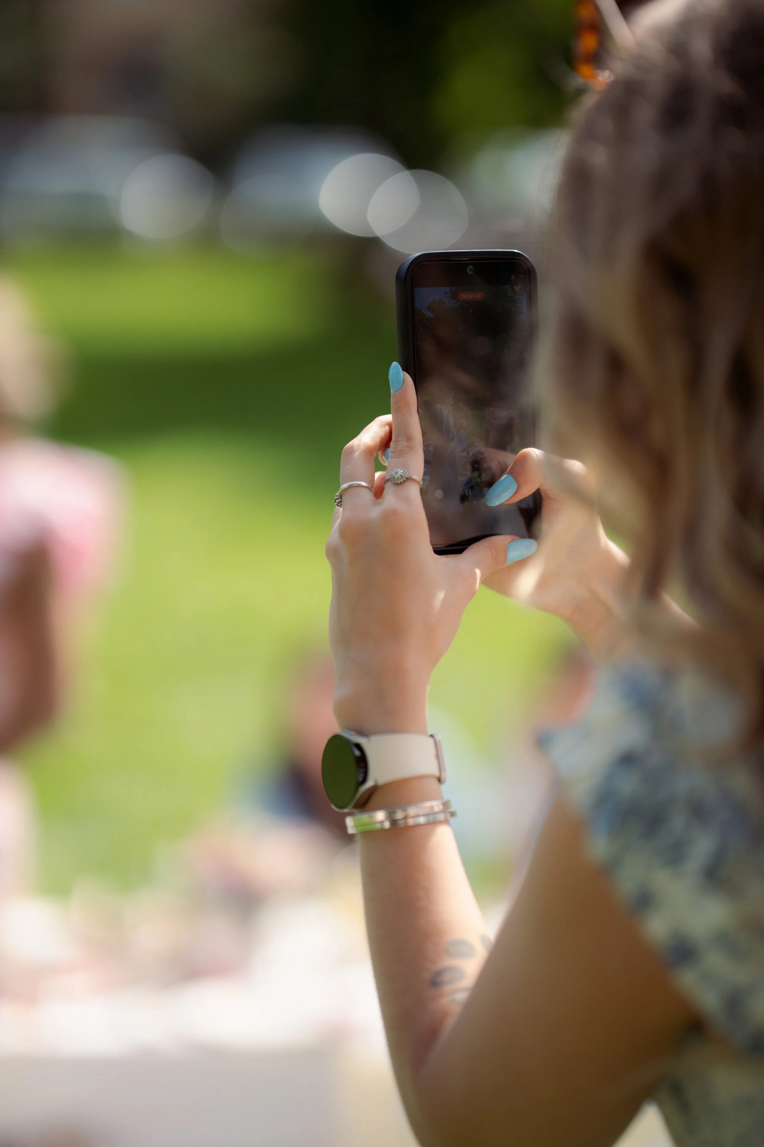 A woman with light blue nails and silver jewelry taking a photo with her smartphone outdoors, with a blurred background of green grass and trees.