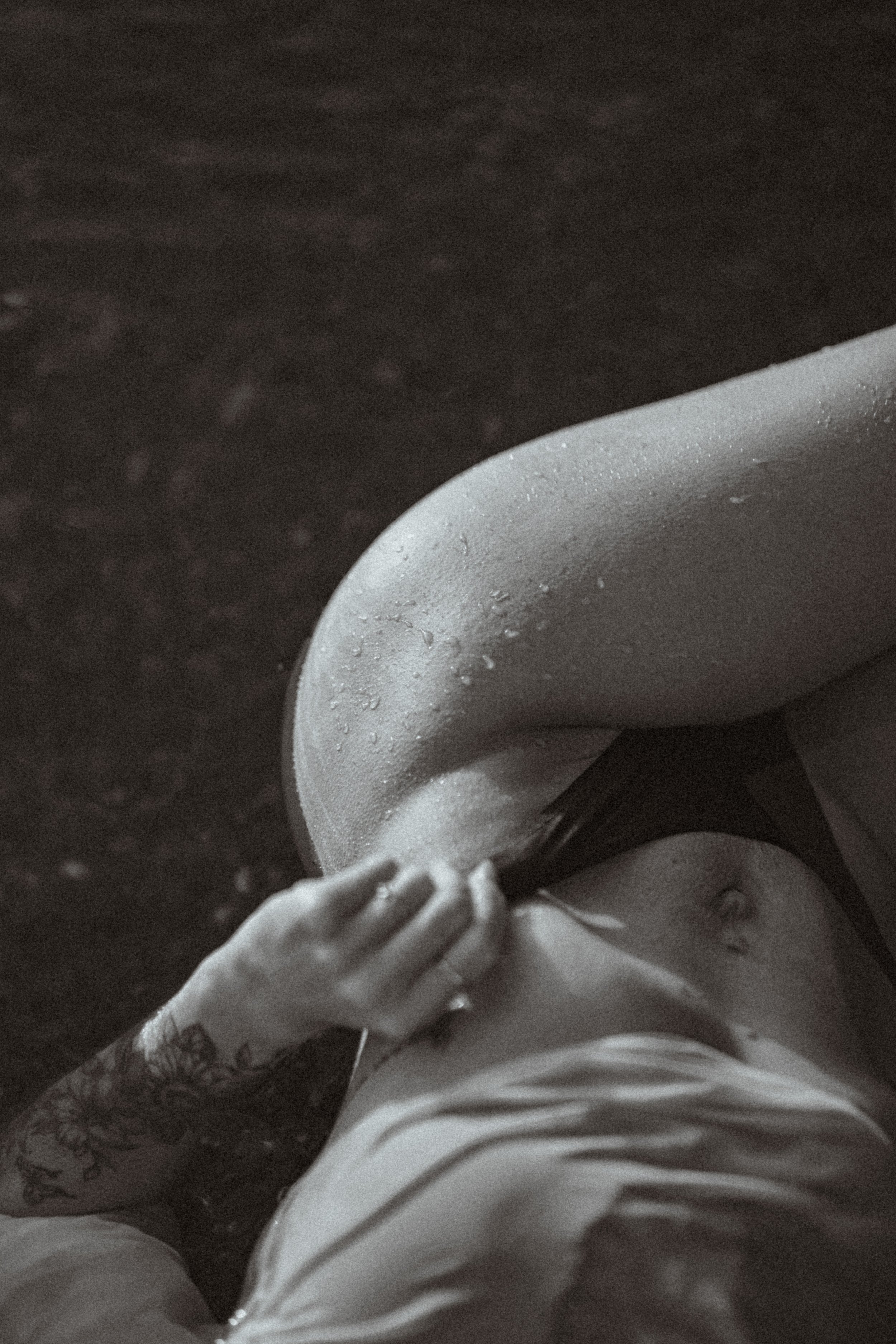 A person lying on their side with droplets of water on their skin, wearing light-colored clothing, with a tattoo visible on their arm, and their hand resting on their chest.