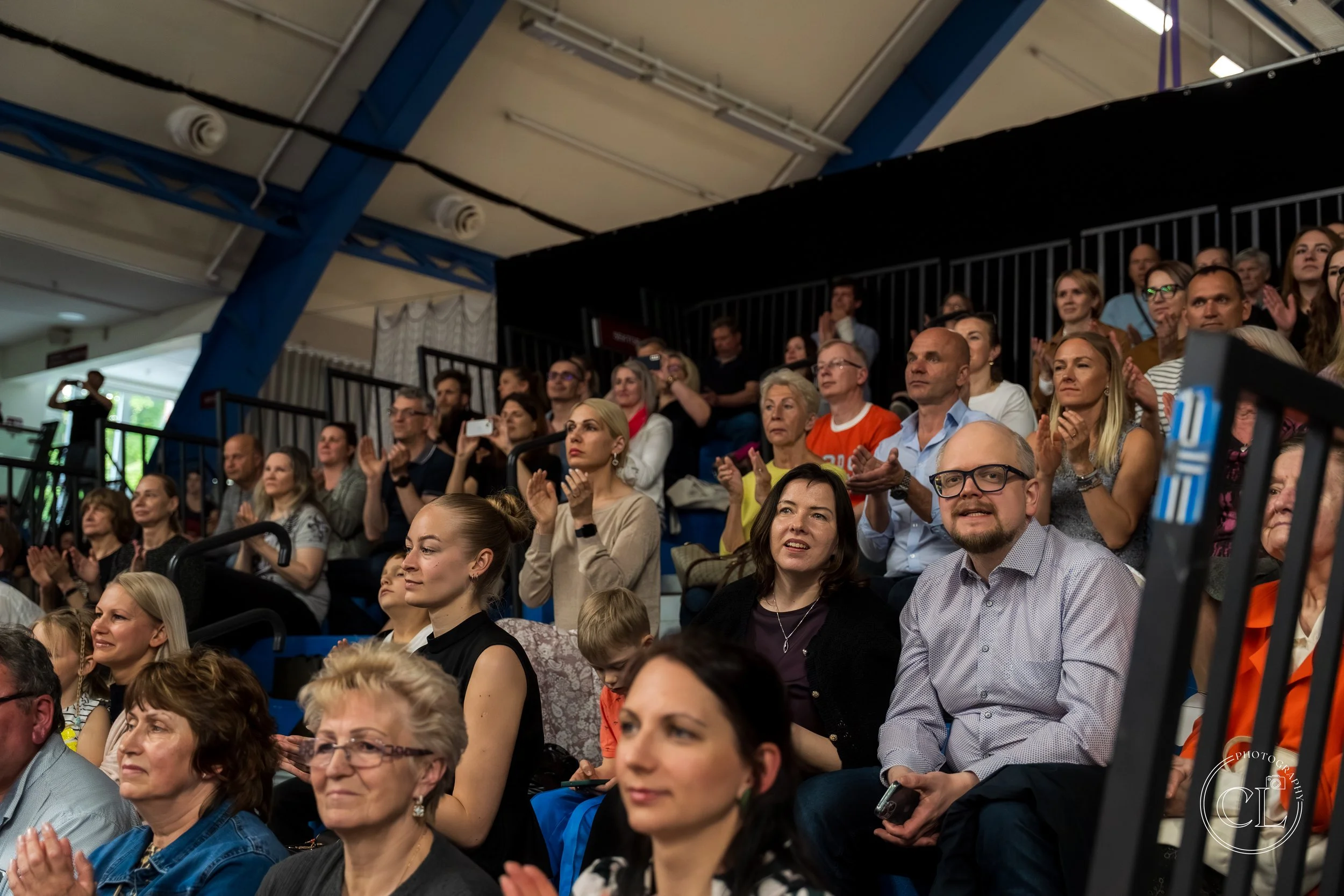 Audience seated in bleachers at a public event, some clapping and listening attentively.
