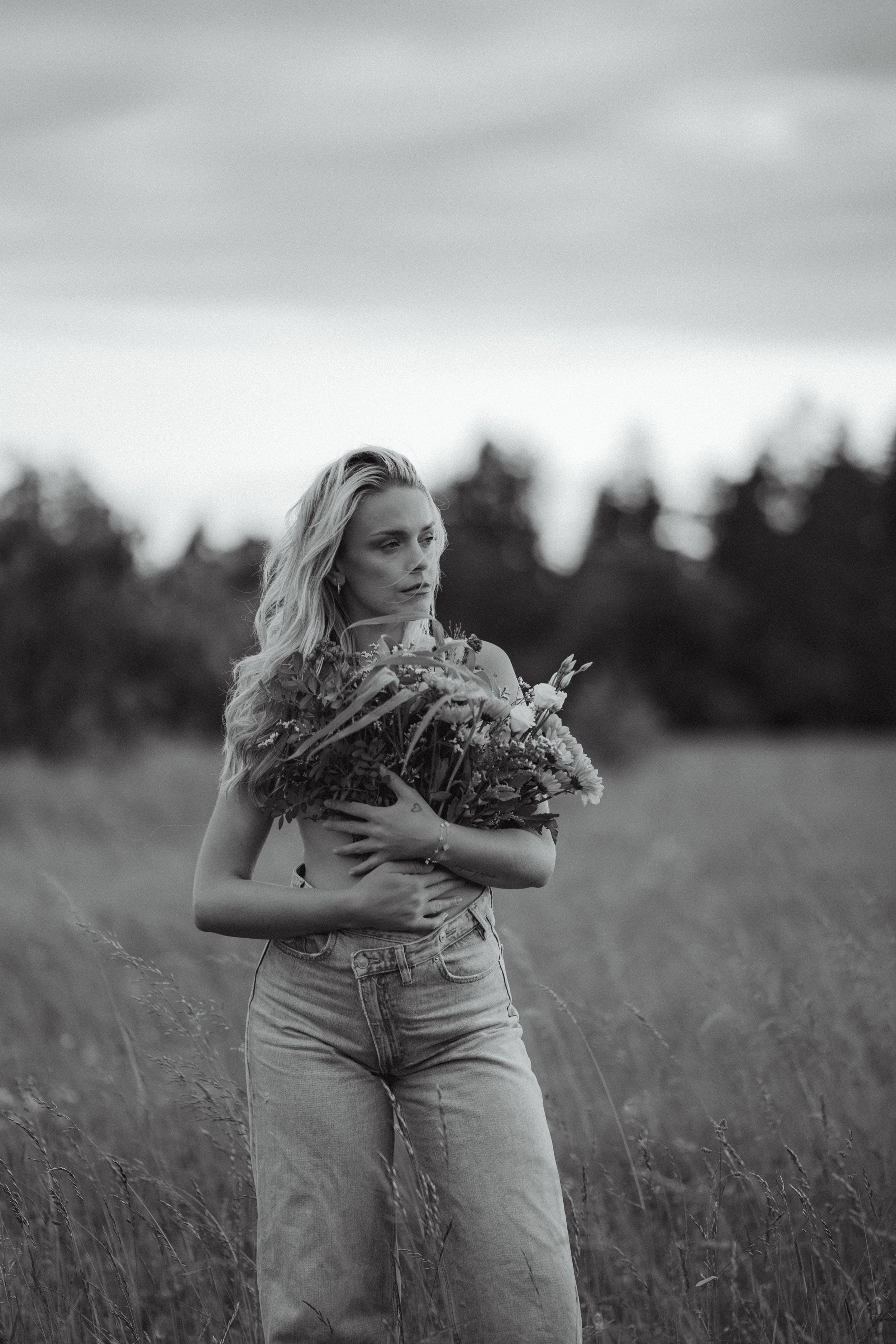 A young woman with long wavy hair standing in a field, holding a bouquet of flowers, gazing into the distance, captured in black and white.