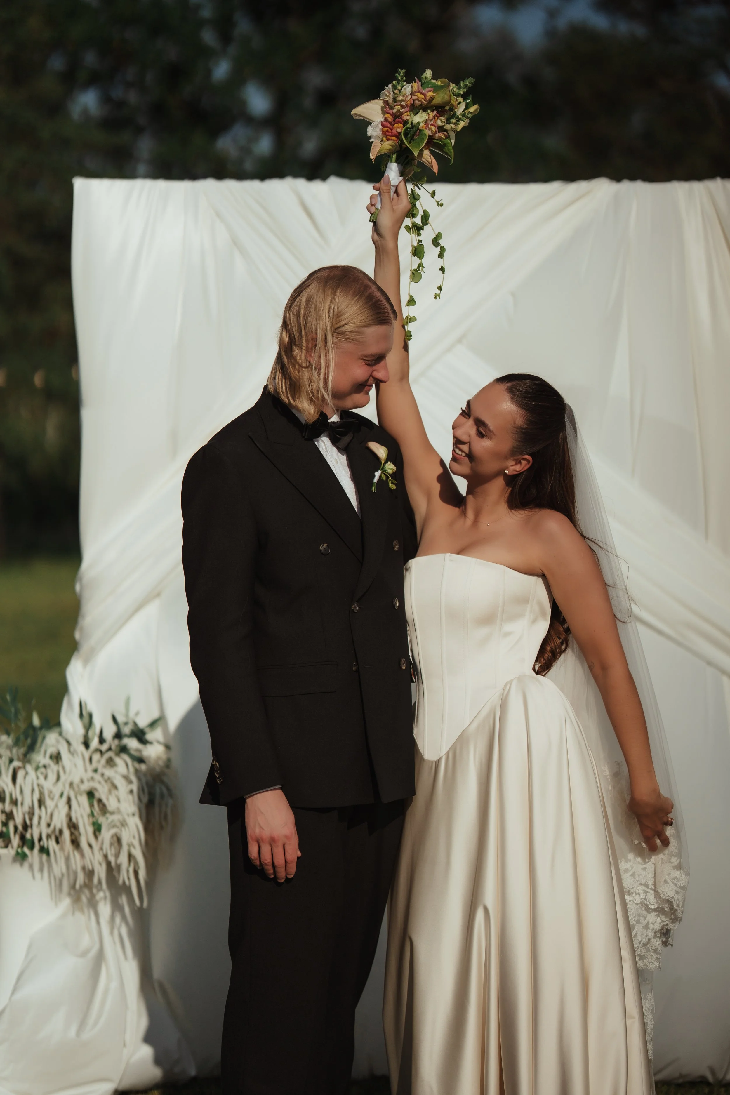 A couple celebrating their wedding outdoors, with the bride holding a bouquet of flowers raised above her head, smiling at each other. The groom wears a black tuxedo and the bride wears a strapless cream-colored wedding dress.