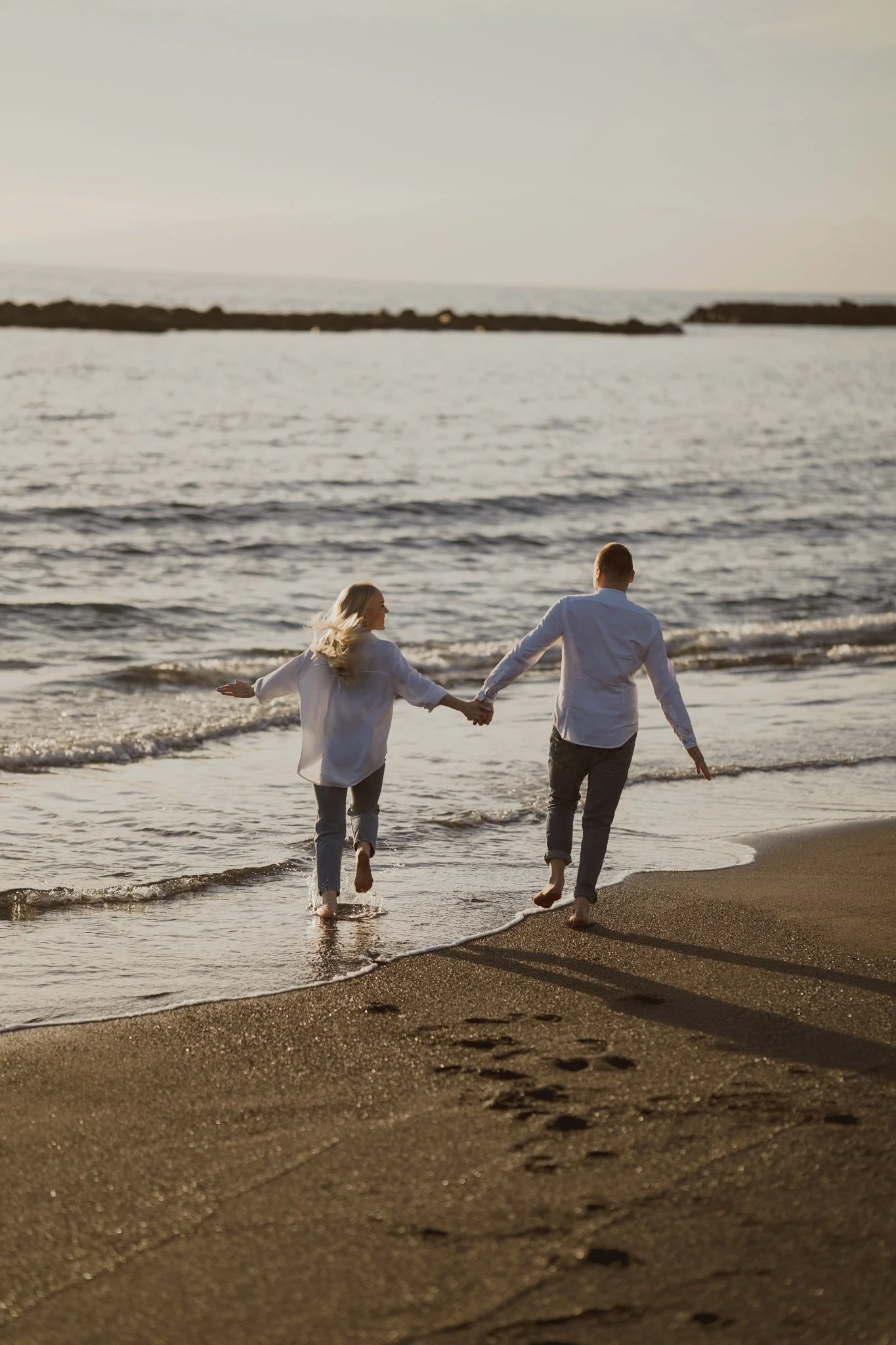 A couple holding hands and running along the shoreline at sunset on the beach.