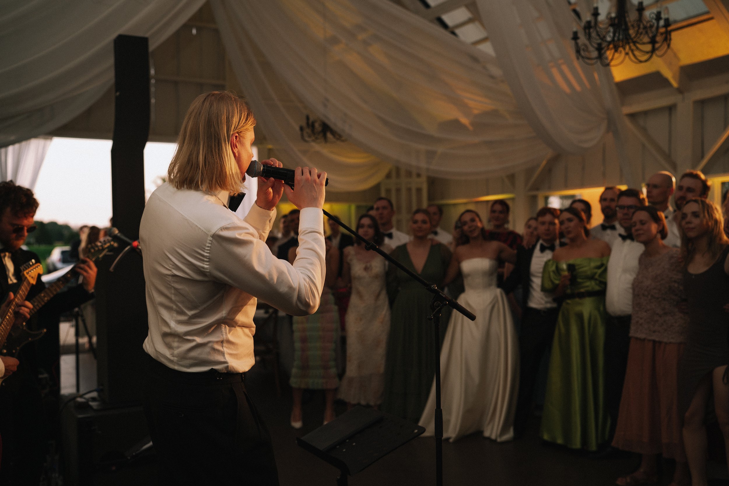 A woman singing into a microphone at a wedding reception with a group of guests watching.