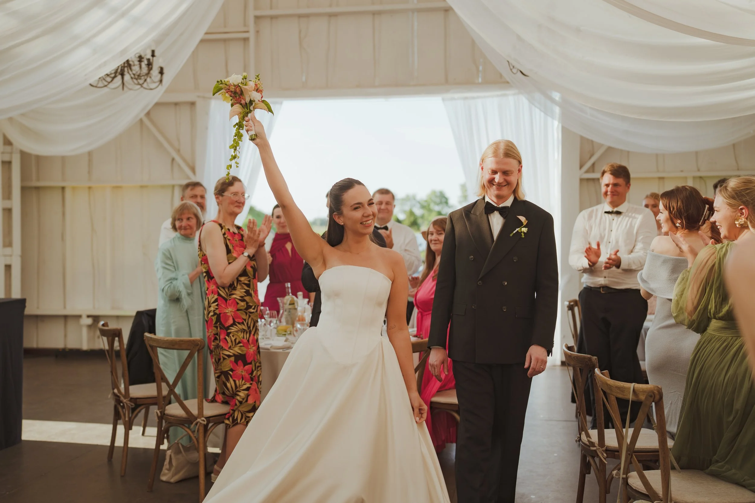 A bride holding a bouquet and smiling, walking with a groom dressed in a tuxedo, as guests applaud in a decorated event space.