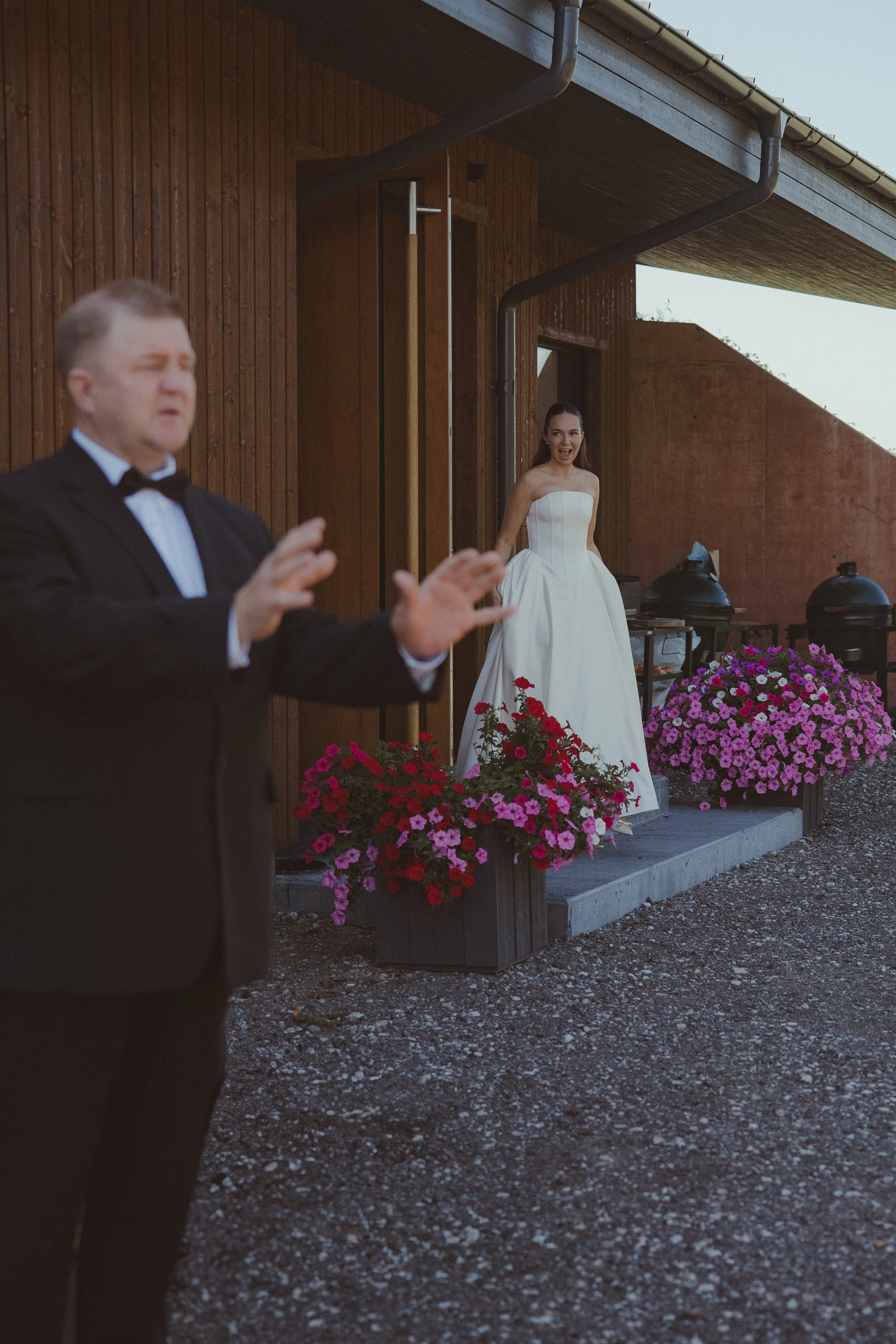 A woman in a white wedding dress reacting happily as a man in a tuxedo gestures in front of her, outside a wooden building with pink and red flowers in planters.