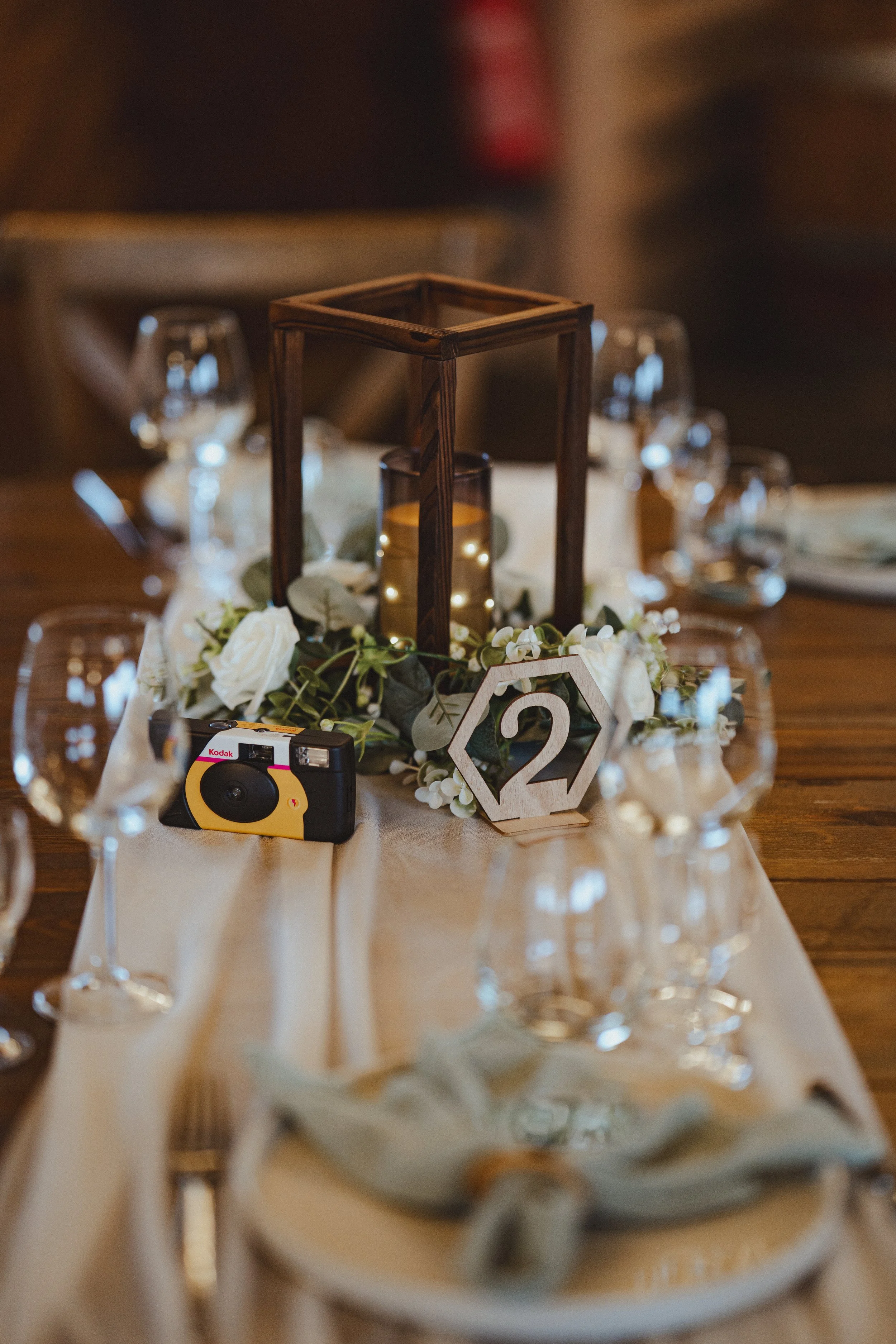Elegant table centerpiece with a lantern, white flowers, a small wooden table number 2, and glassware.