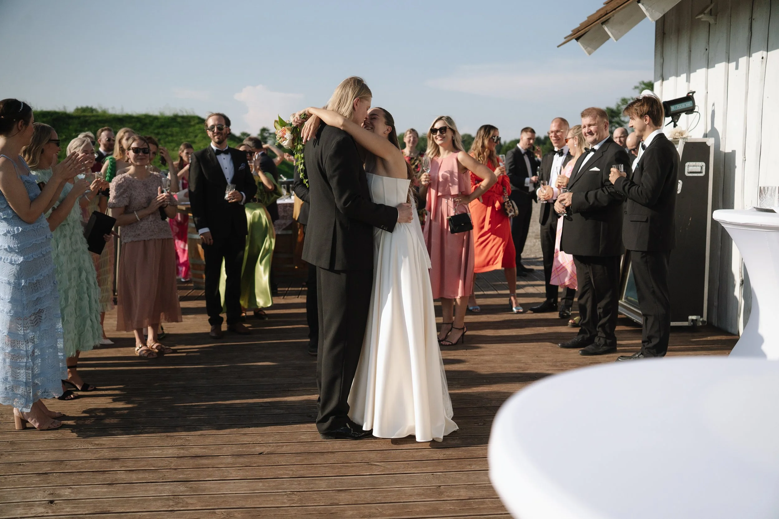 Bride and groom dancing at their wedding reception held outdoors on a wooden deck. Guests in formal attire are watching and celebrating the couple.