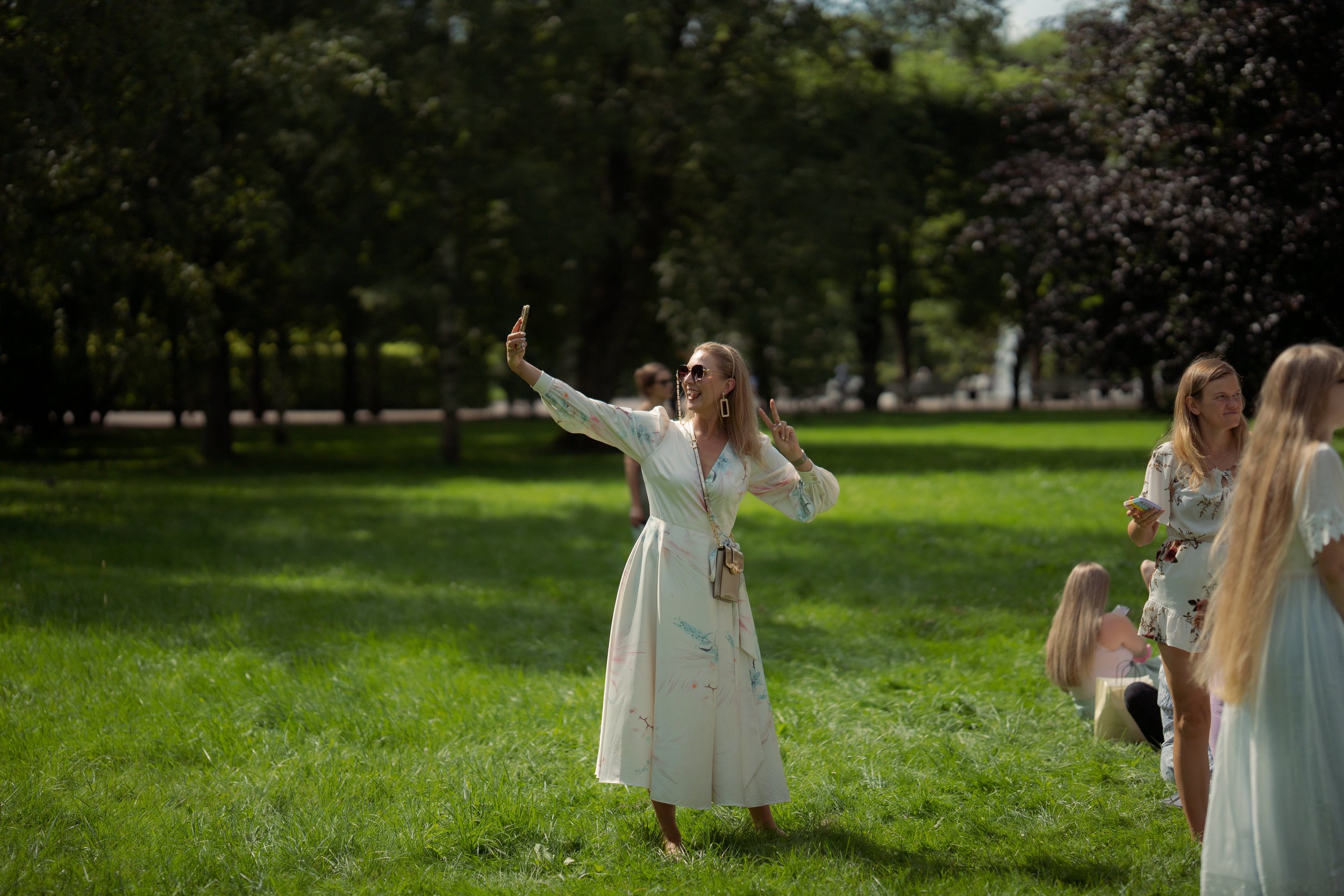 A woman in a long, light-colored dress taking a selfie in a park with green grass and trees.