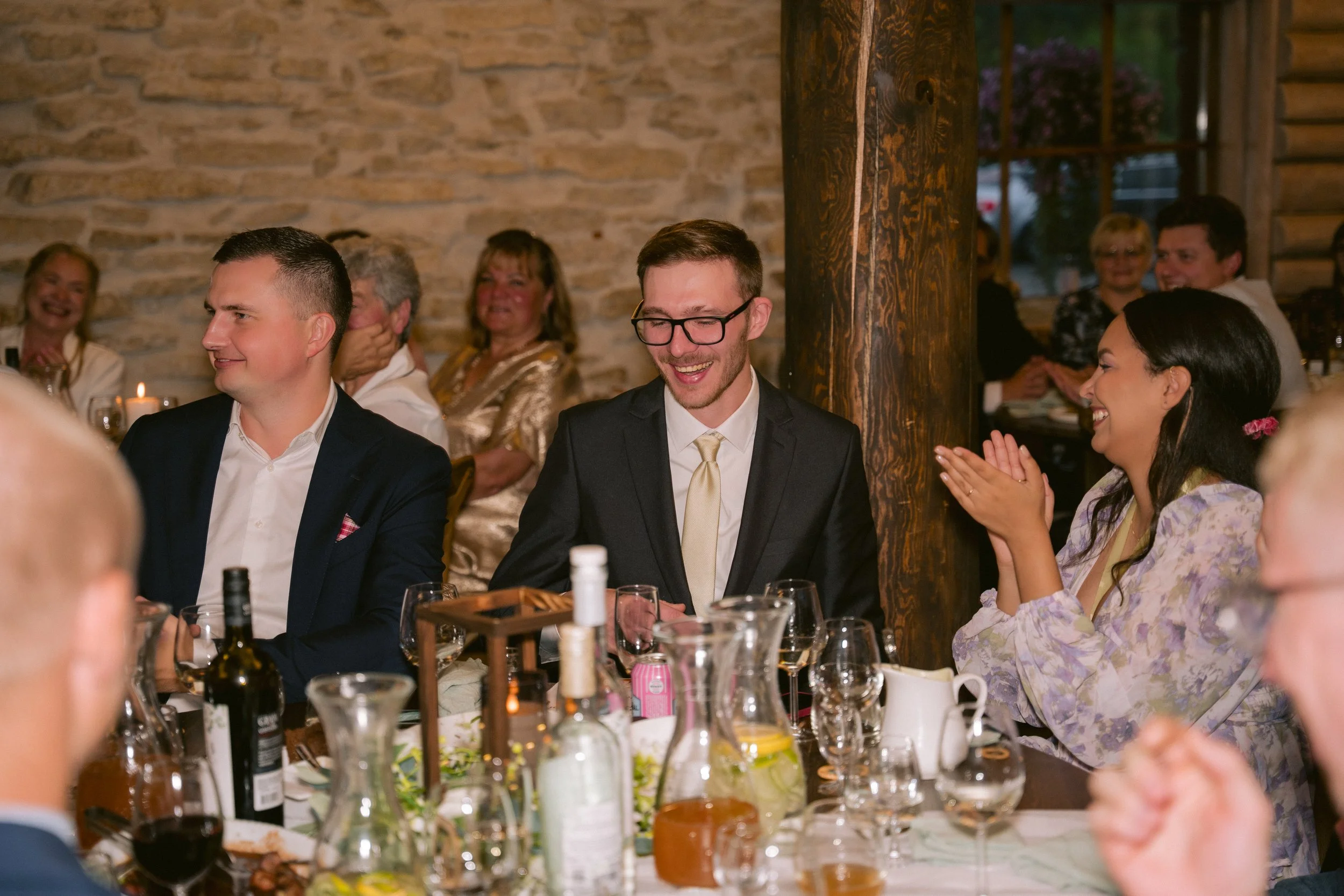 A group of people at a celebration or wedding reception, sitting at a table with drinks, smiling, and clapping, in a rustic venue with stone walls and wooden beams.