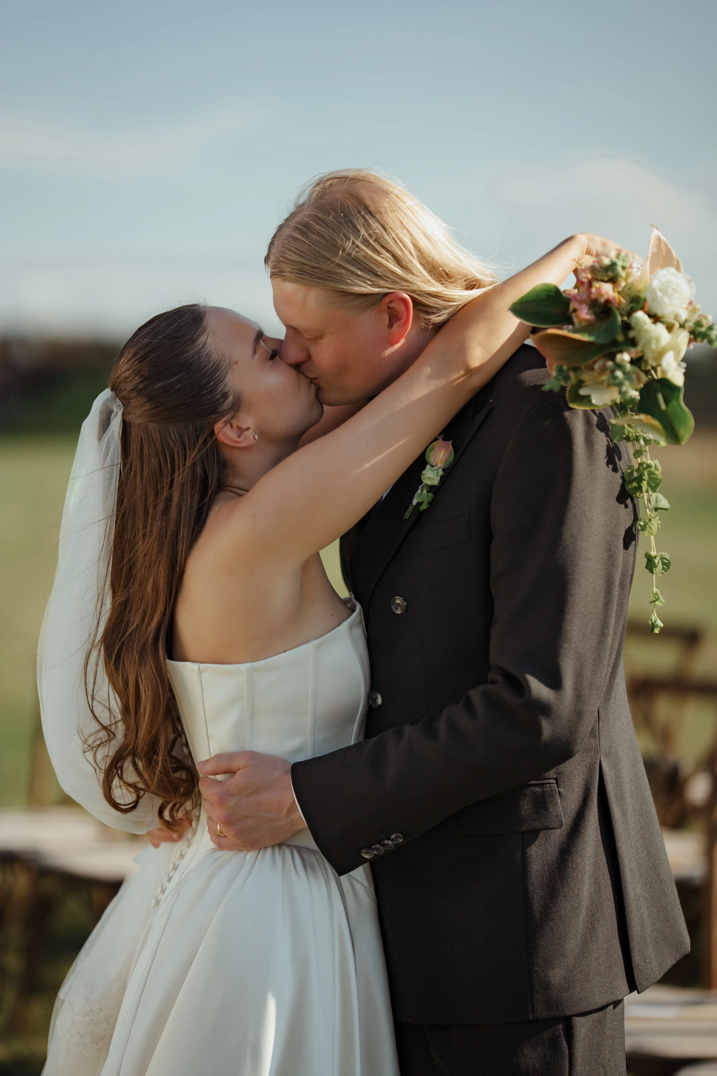 A bride and groom sharing a kiss outdoors on their wedding day, with the bride holding a bouquet of flowers.