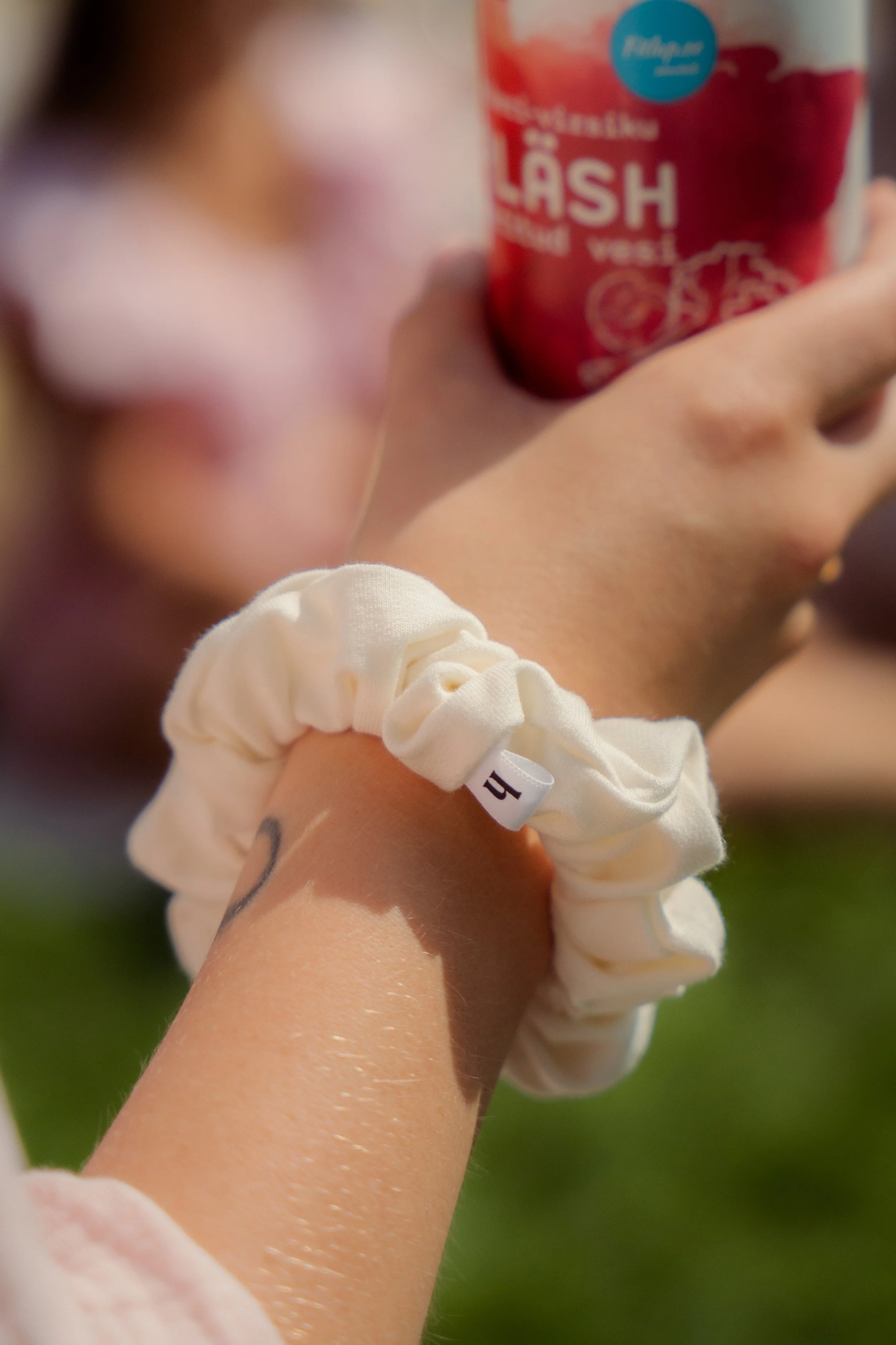 Close-up of a person's arm with a cream-colored scrunchie and a tattoo, holding a container of cherry-flavored chewing gum, with a blurred face of someone in the background.