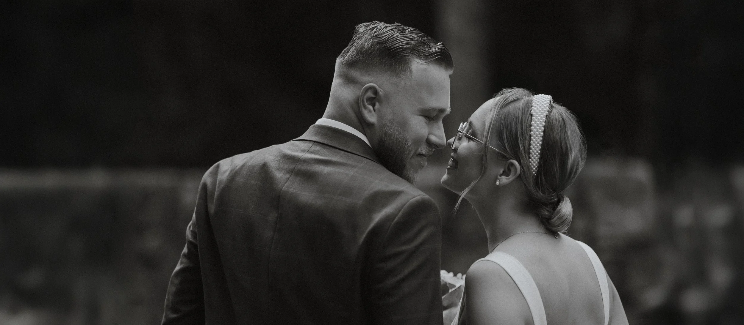 A black and white photo of a man and woman dressed in wedding attire, standing close and gazing into each other's eyes, smiling.