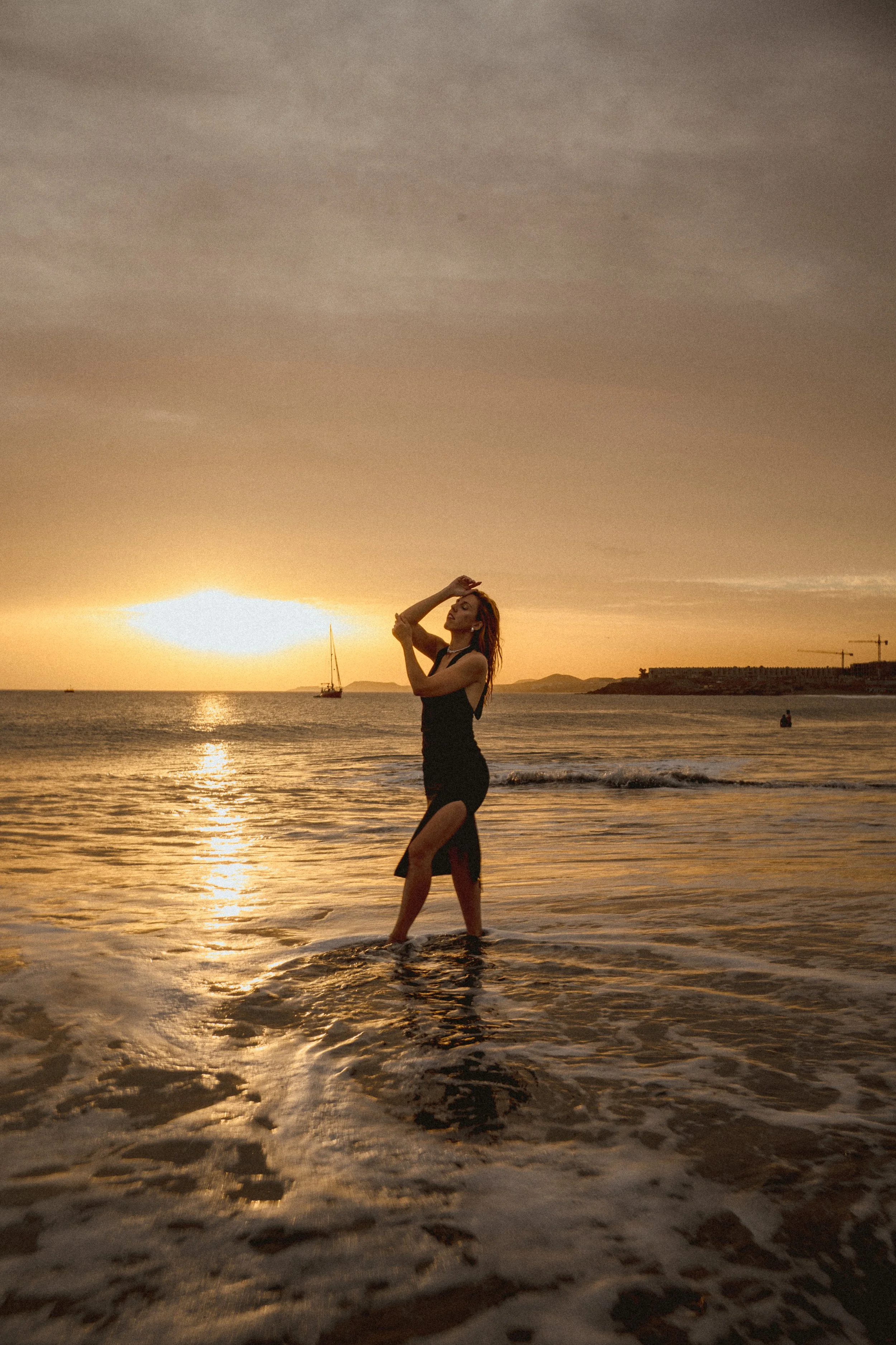 A woman standing in the water at sunset, with her hands raised near her face, on a beach with sailboats and distant land in the background.