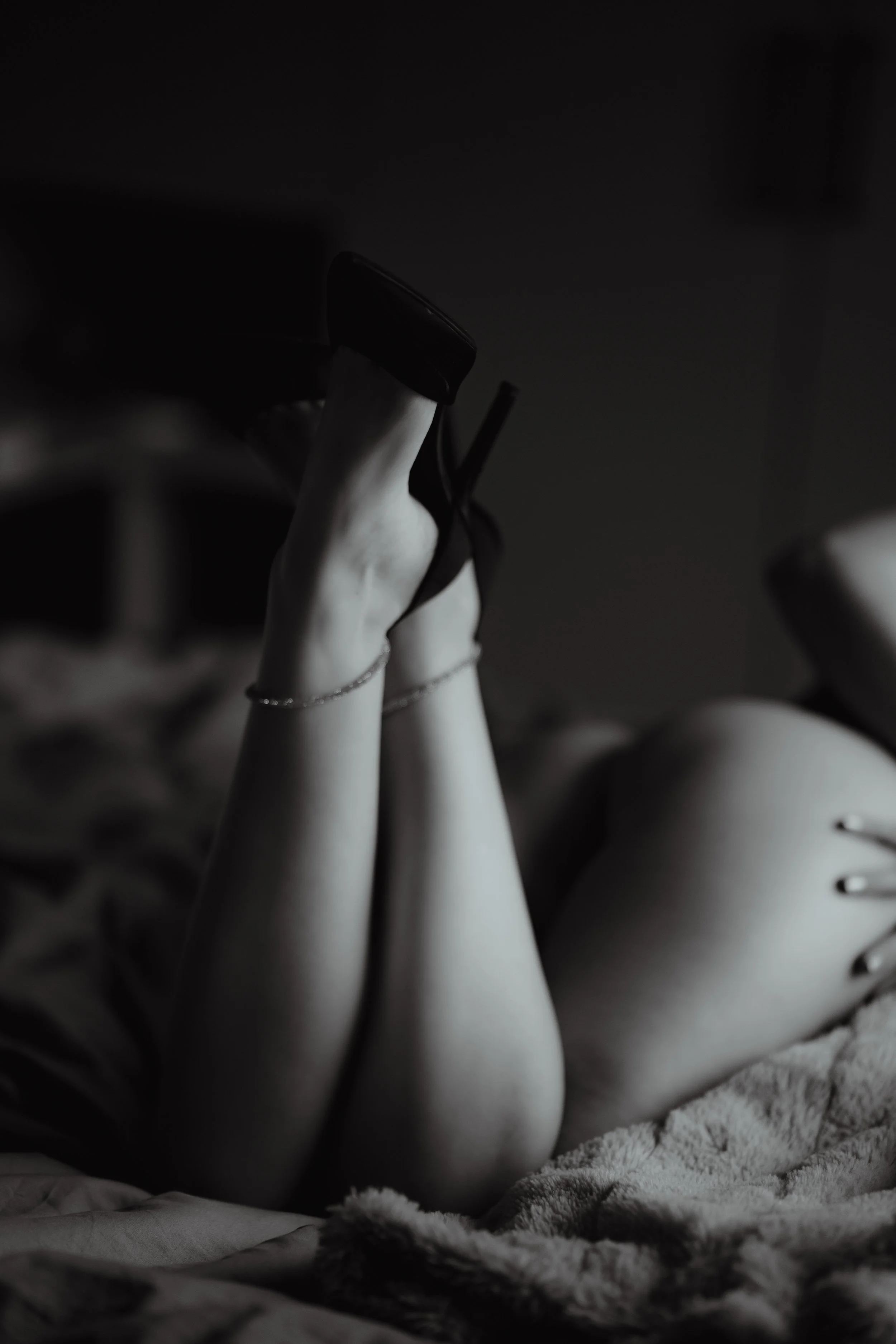 Black and white photo of someone's legs and feet raised in the air while lying on a bed, with the person wearing high heels and anklets.