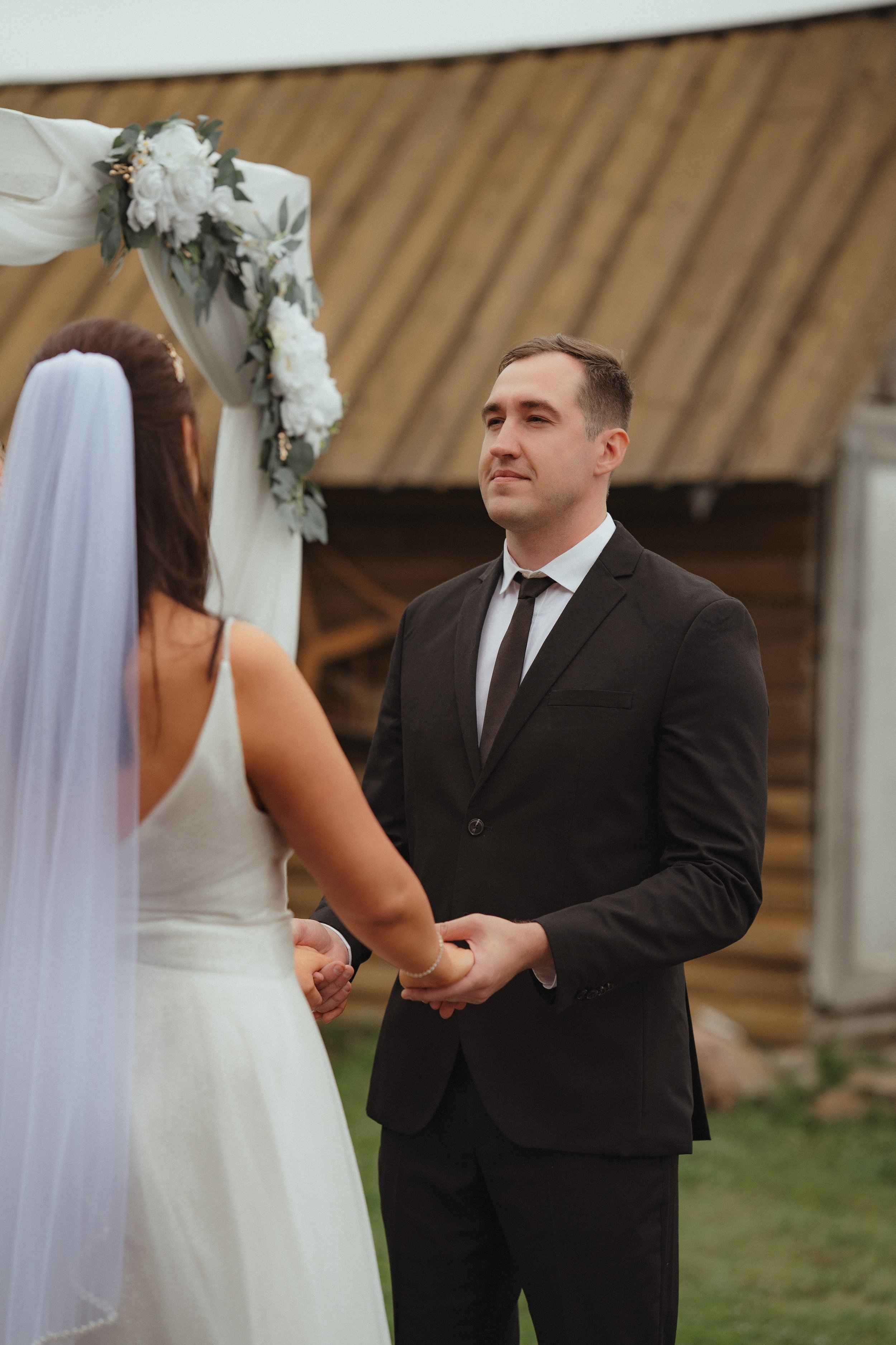 A groom in a black suit and white shirt holding hands with a bride in a white wedding dress, during their outdoor wedding ceremony, with a wooden building and floral wedding arch visible in the background.