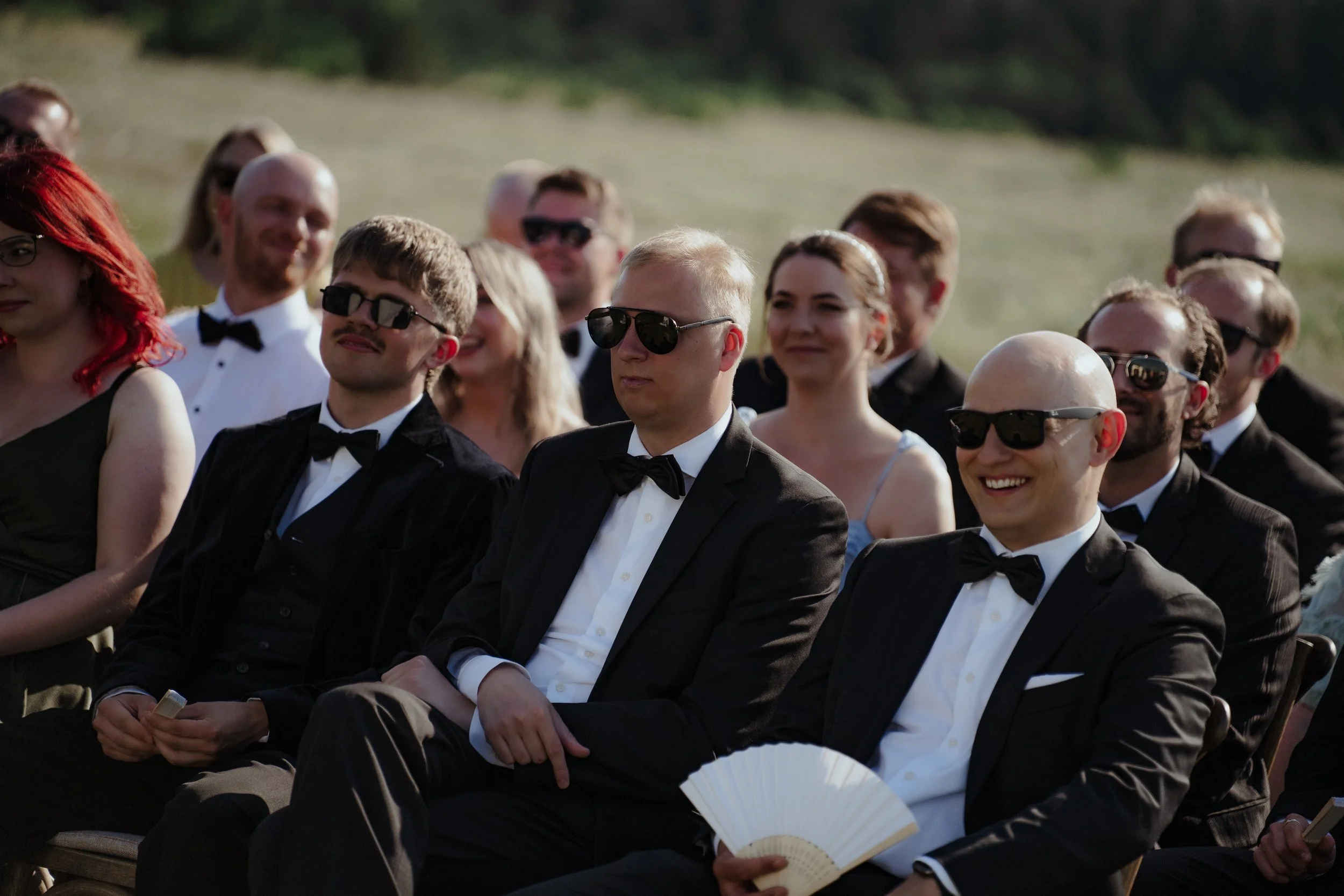 A group of people dressed in formal attire, sitting outdoors, many wearing sunglasses, during a daytime event.