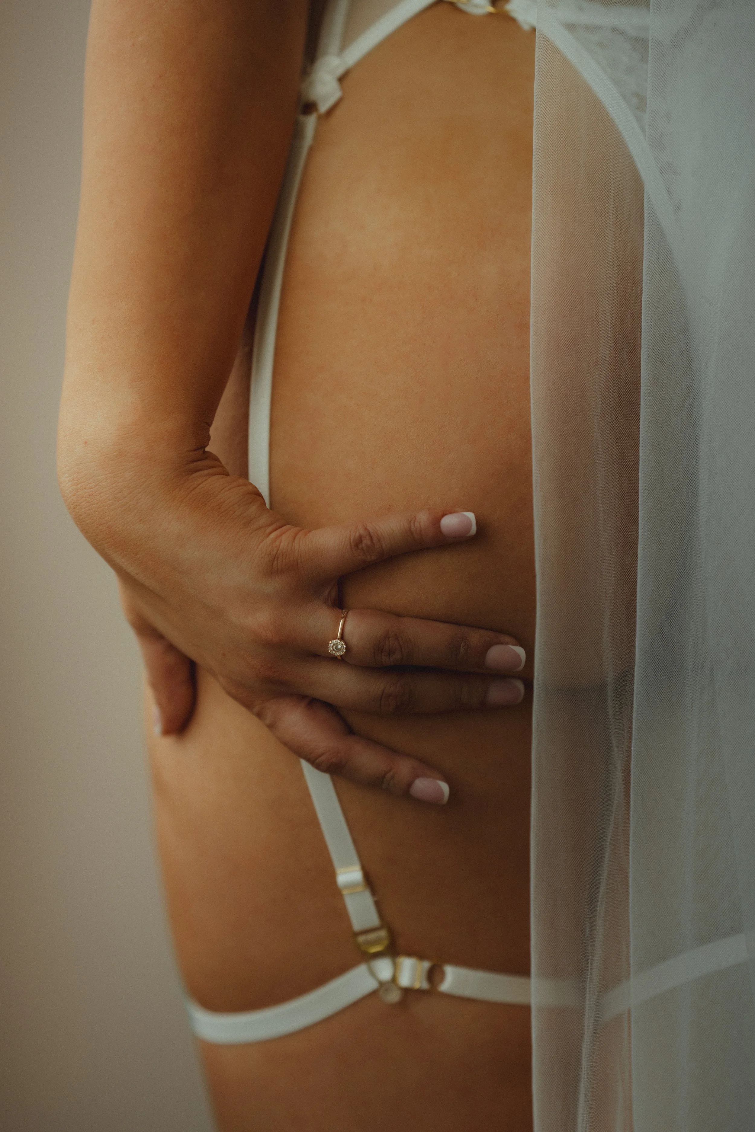 Close-up of a woman’s hand with a ring on her finger resting on her thigh, wearing white lingerie and a sheer garment.