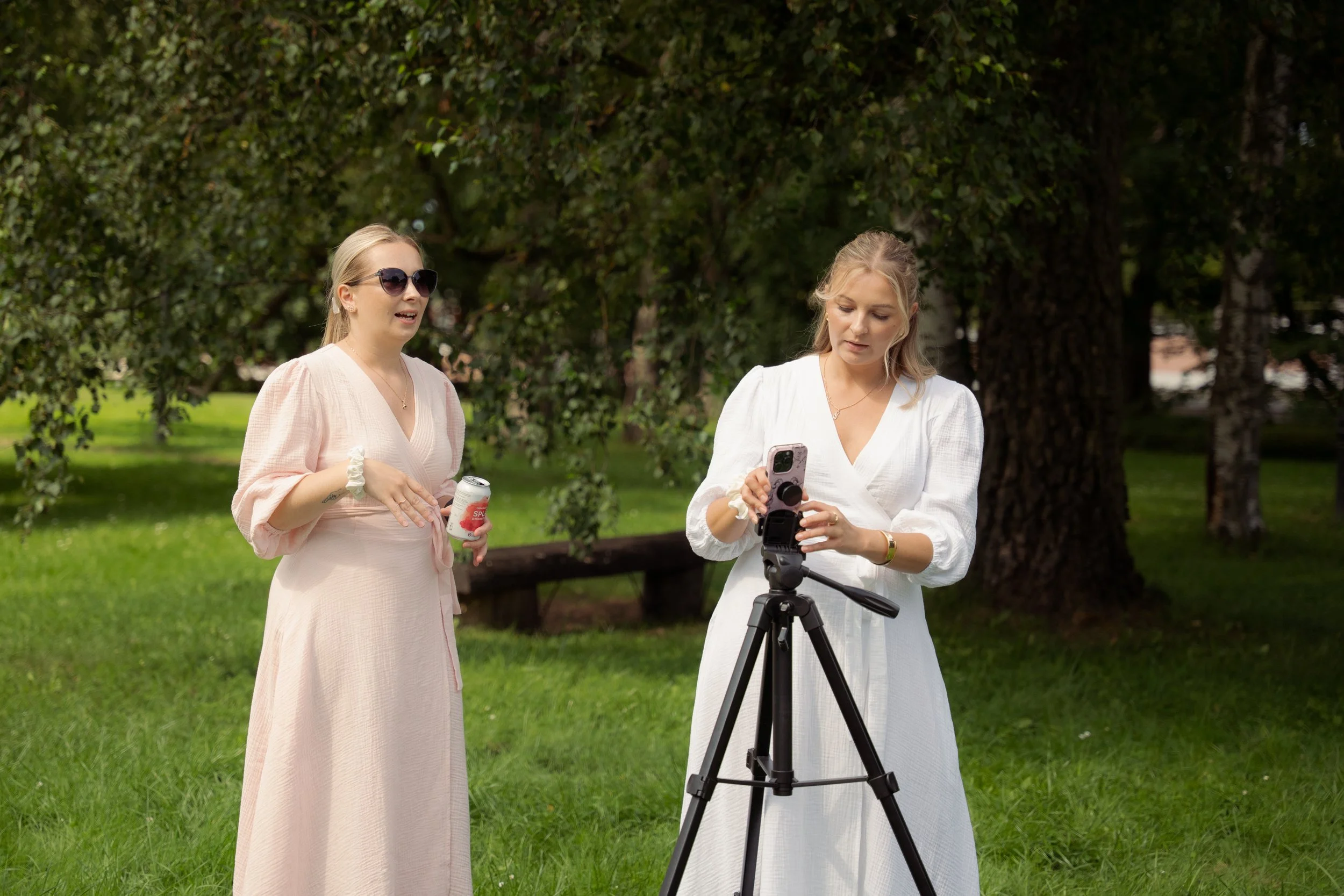 Two women standing outdoors in a park with green grass and trees, one adjusting a smartphone on a tripod while the other holds a soda can and looks on.