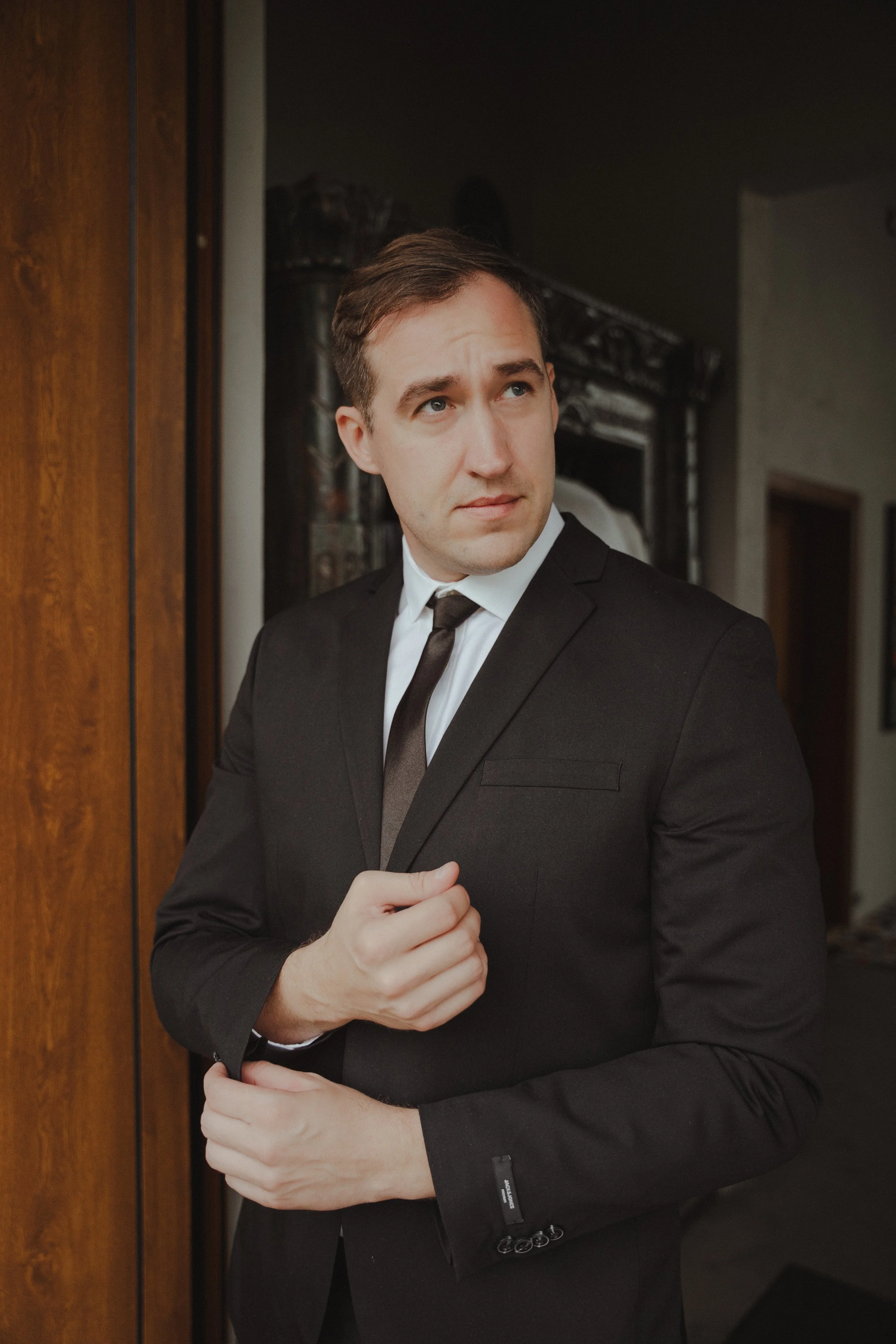 A young man in a black suit and tie standing indoors, adjusting his suit jacket, with a thoughtful expression.