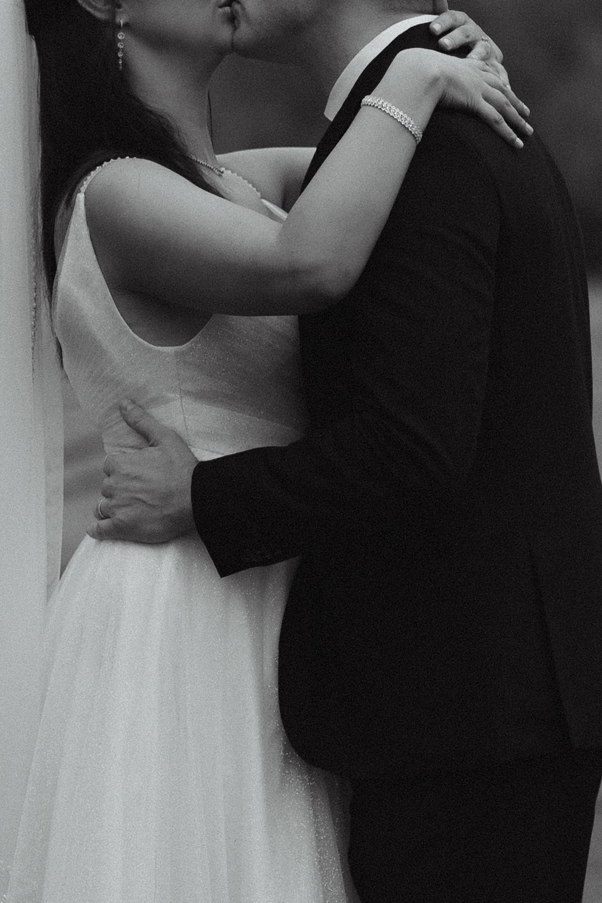 A black and white photo of a couple kissing tightly, the woman wearing a sleeveless wedding dress with fine details, jewelry, and the man in a dark suit, holding each other closely.
