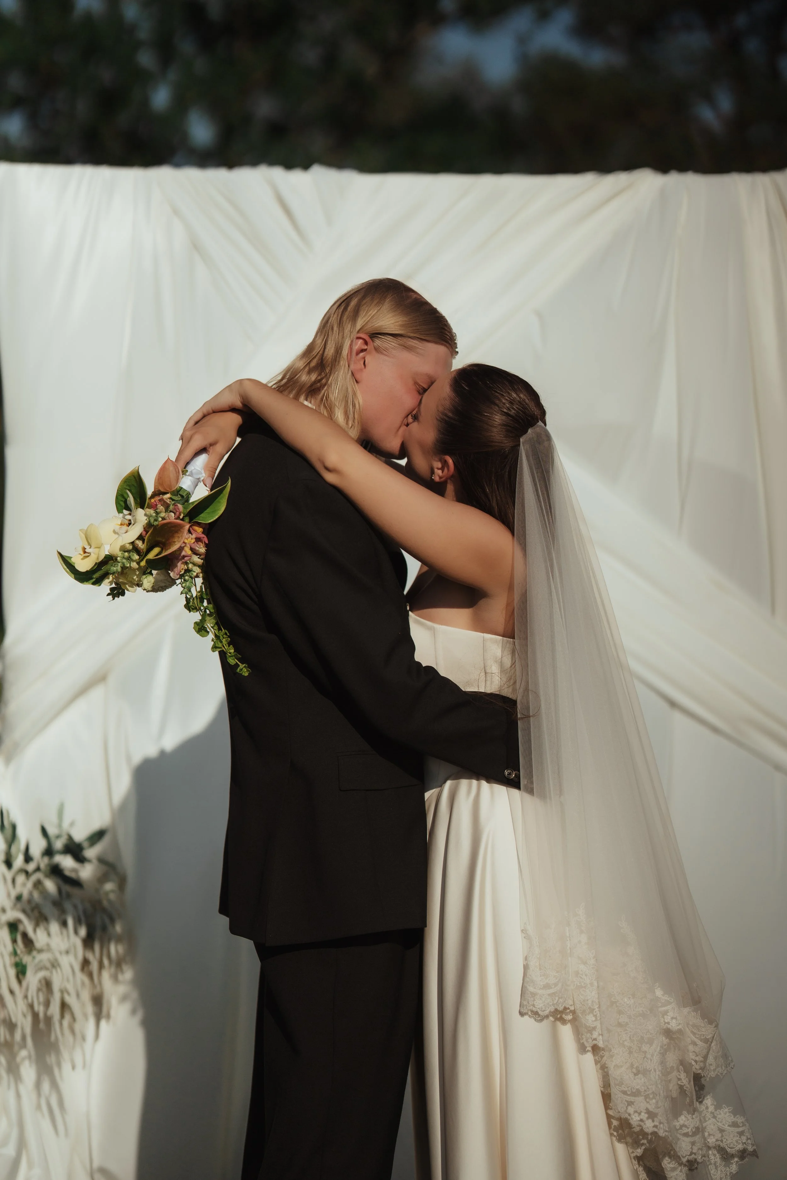 Two brides, one in a black suit and the other in a white wedding dress, kissing during a wedding ceremony with a white draped backdrop.