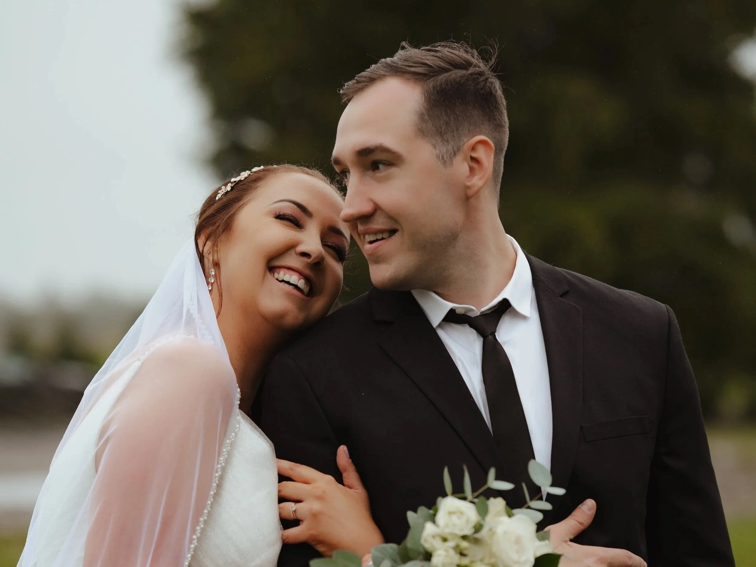 A smiling bride and groom in wedding attire outdoors, with the bride leaning her head on the groom's shoulder, holding a bouquet of white flowers, in a joyful moment.