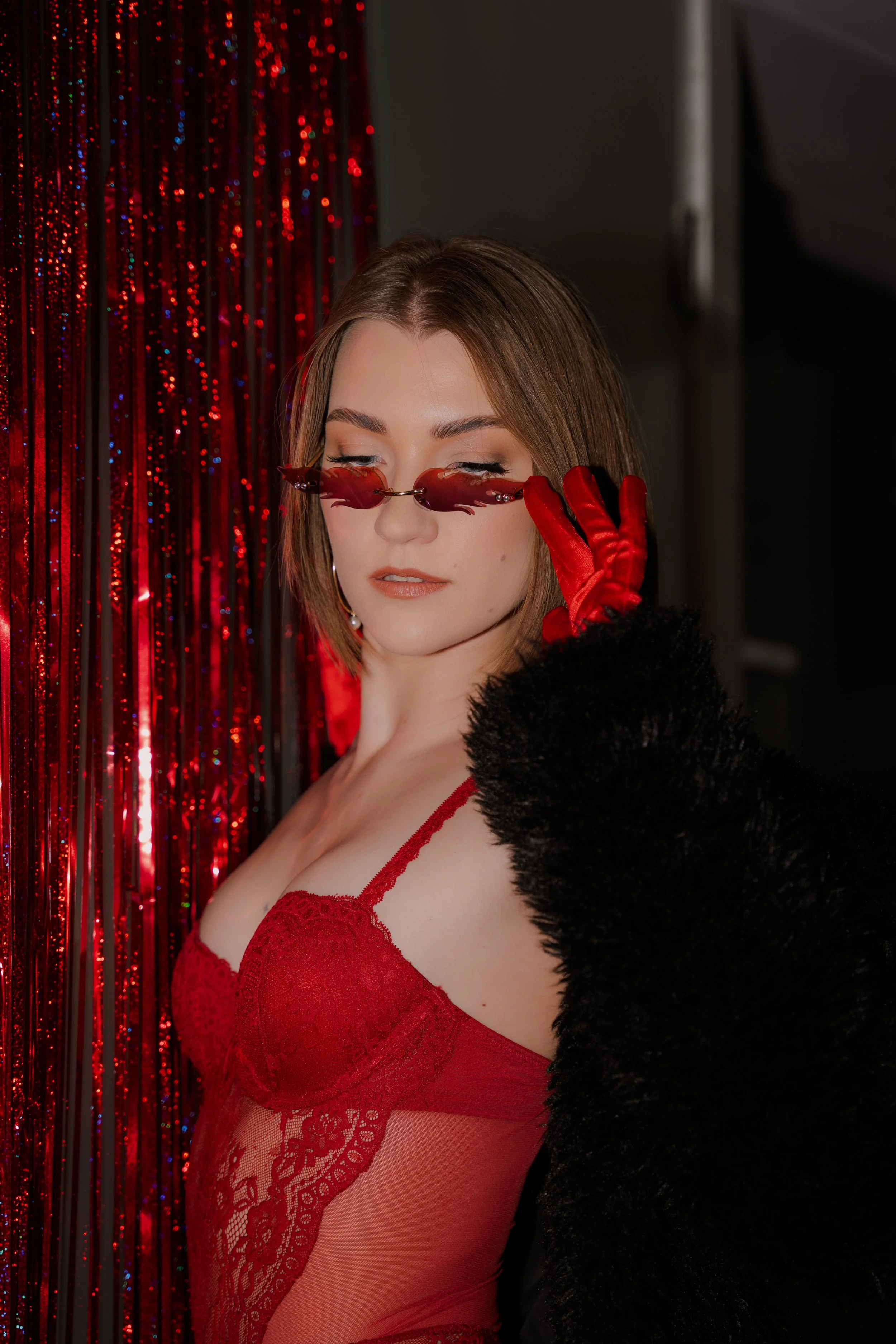 A woman with short brown hair is wearing red lace lingerie, red gloves, and unique red sunglasses. She is standing in front of a red, sparkly curtain, striking a confident pose with her eyes closed and lips slightly parted.