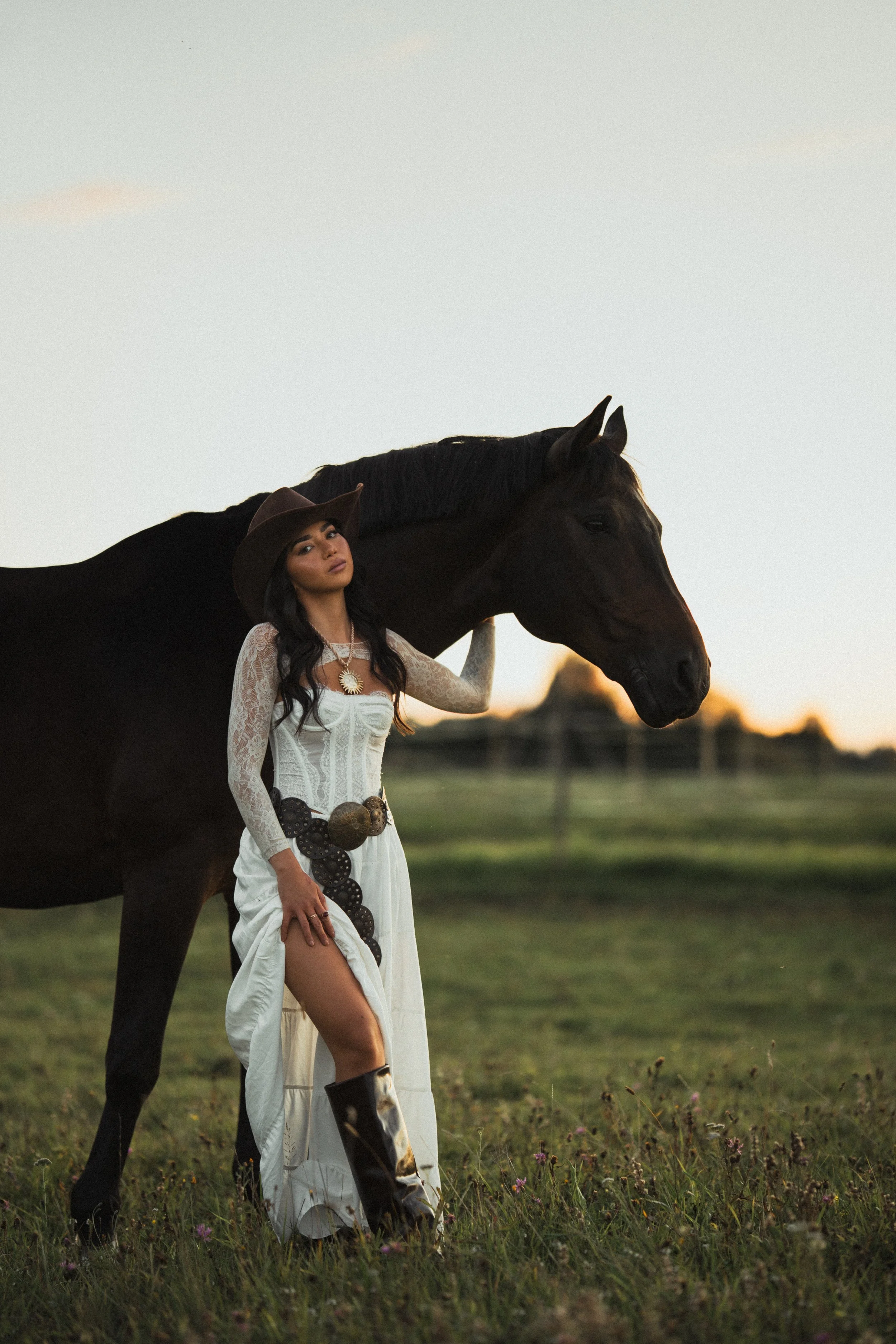 A woman in a white dress with lace sleeves and a large belt, wearing a cowboy hat and black cowboy boots, stands next to a black horse in a grassy field at sunset.