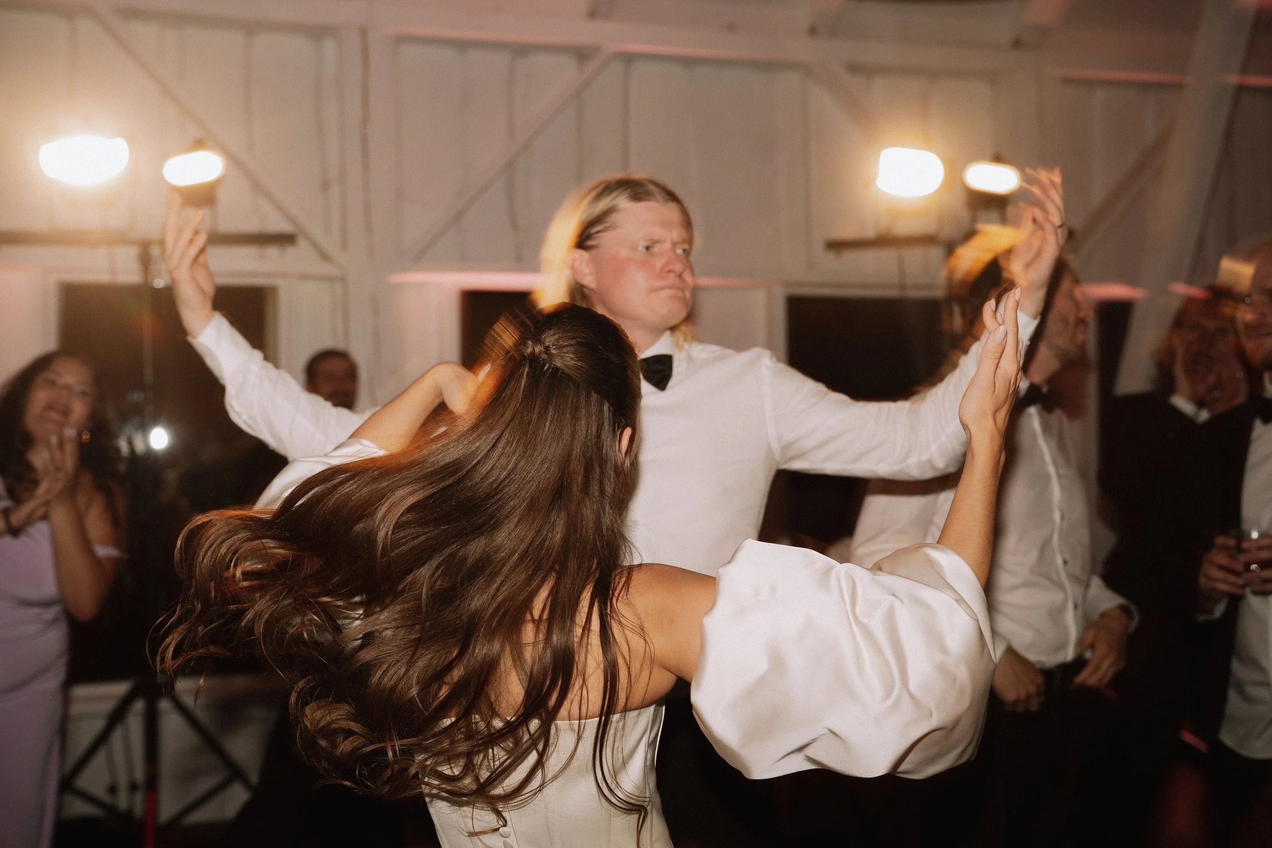 People dancing and celebrating at a party with warm lighting and a wooden ceiling.