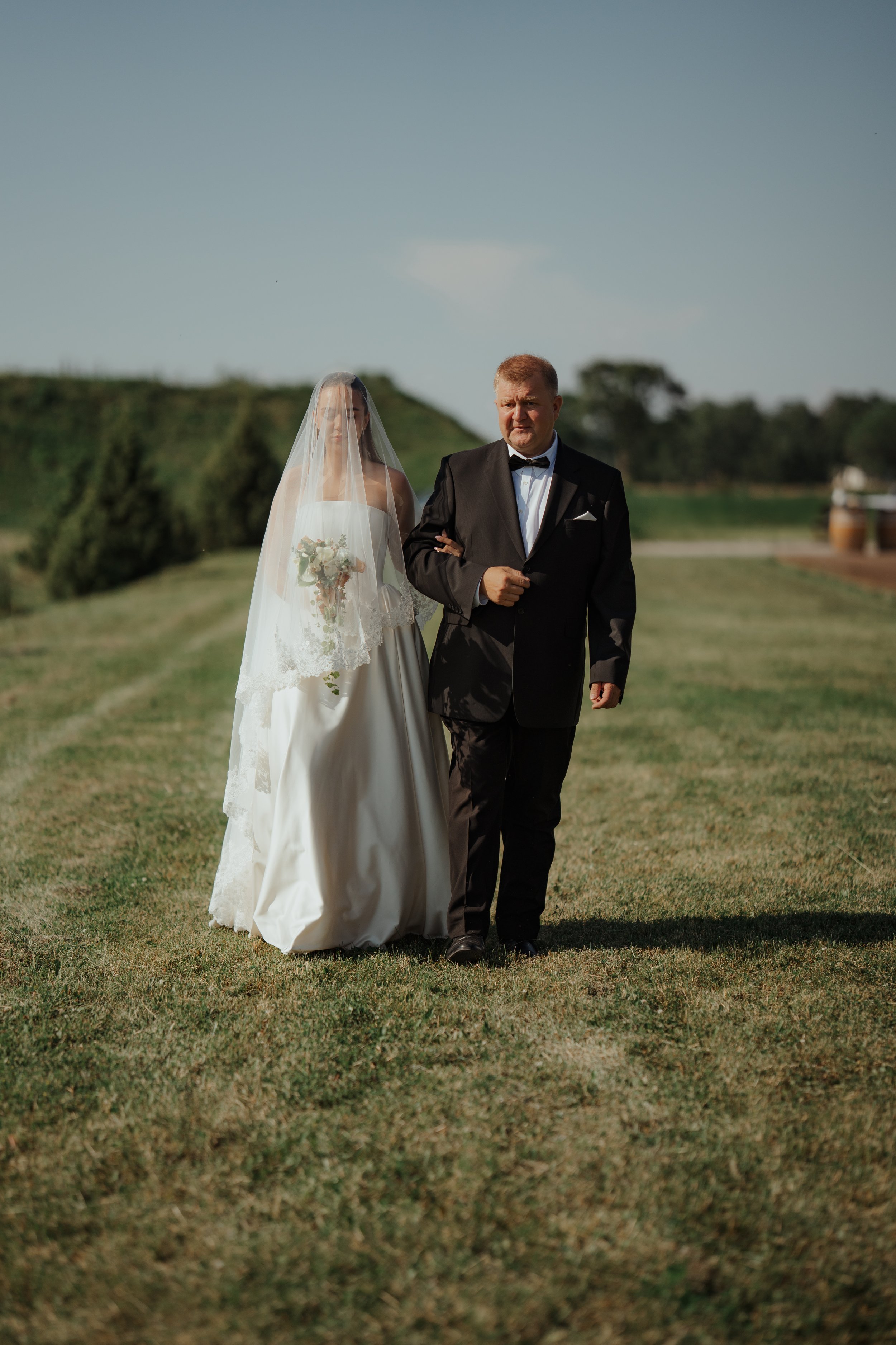 A bride walking down the aisle with a man, possibly her father, outdoors on a grassy field under a clear blue sky.