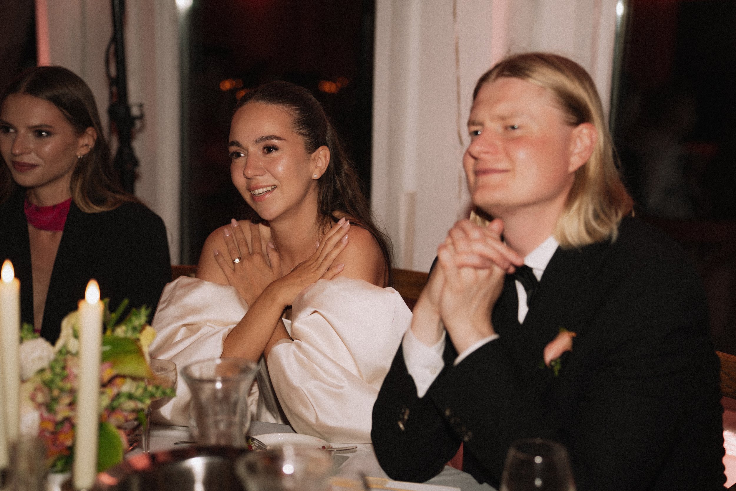 People attending a formal dinner; woman in a white dress smiling, man in a tuxedo, woman in black attire sitting beside her.