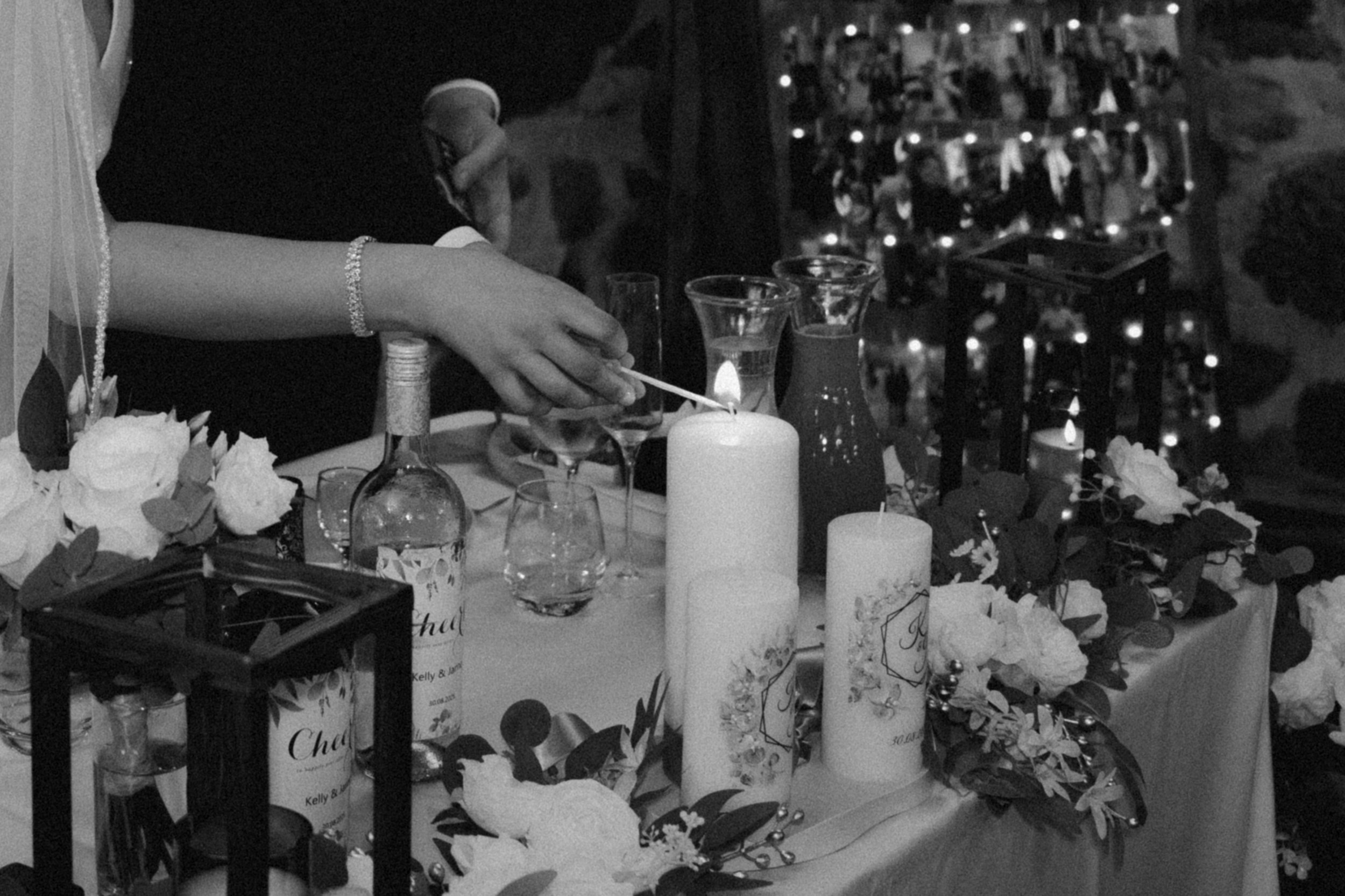 A person lighting a white candle on a decorated table with flowers, candles, and bottles, likely at a celebration or event in a dimly lit setting.
