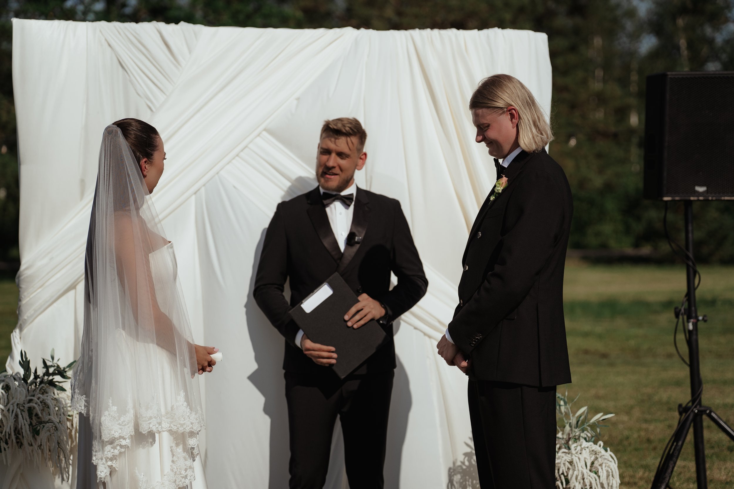 Bride and groom standing outdoors during wedding ceremony, officiant speaking, with white backdrop and speaker nearby.