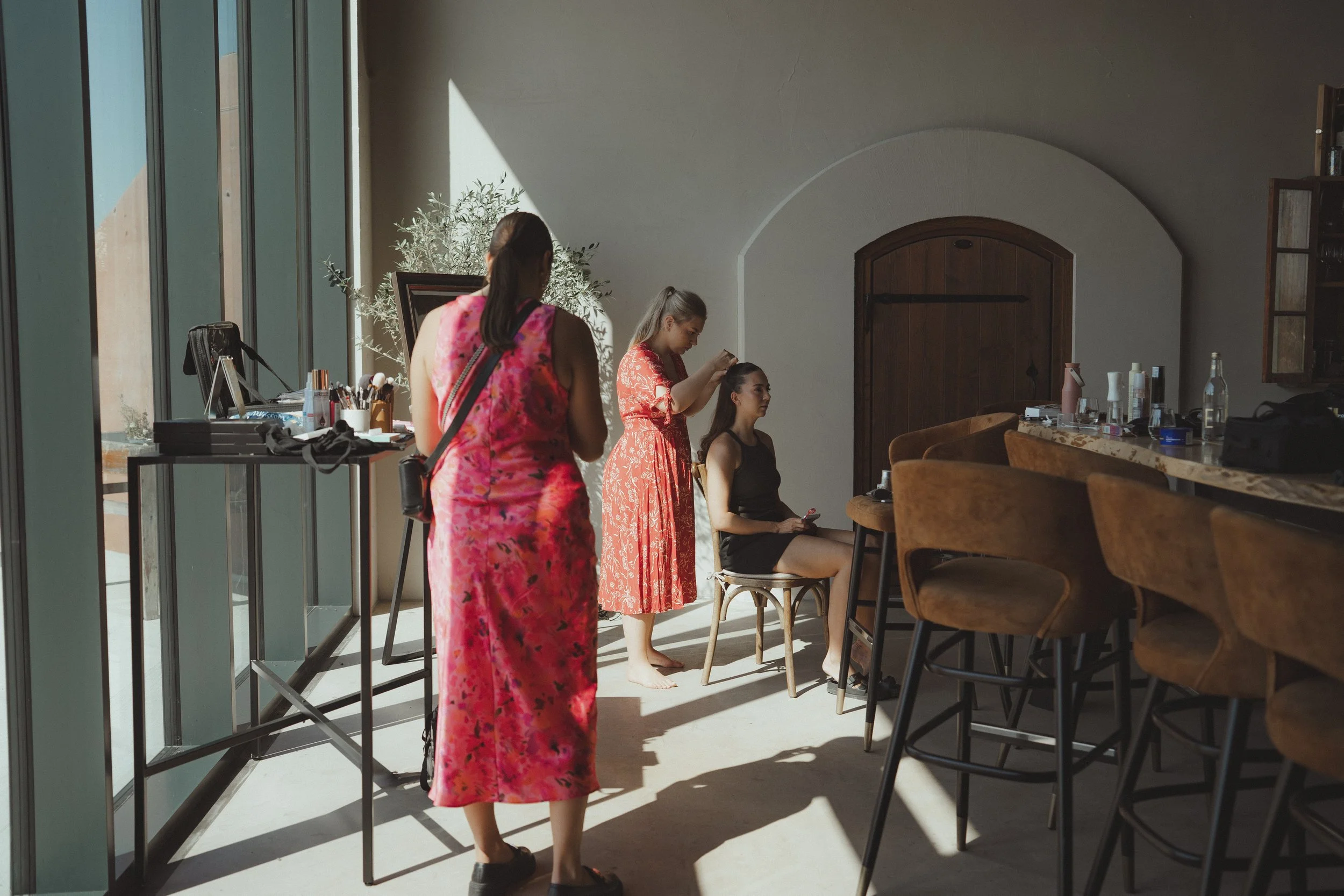 A woman in a black dress is getting her hair styled by a hairstylist in a red dress. Another woman in a pink floral dress is standing nearby. The scene takes place in a well-lit room with large windows, wooden furniture, and a bar counter with variou