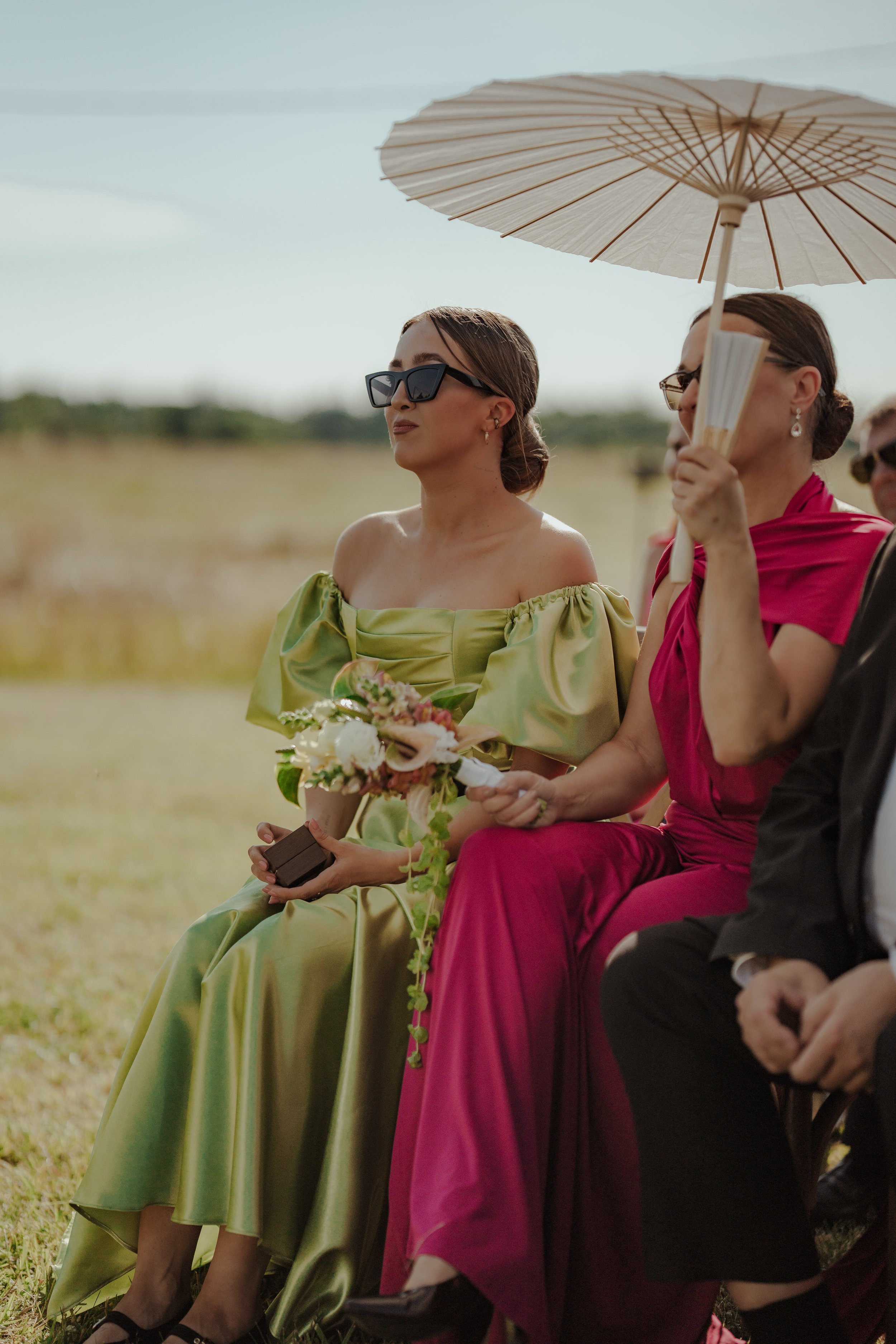 Women dressed in colorful dresses sitting outdoors at a daytime event, with one woman holding a parasol.