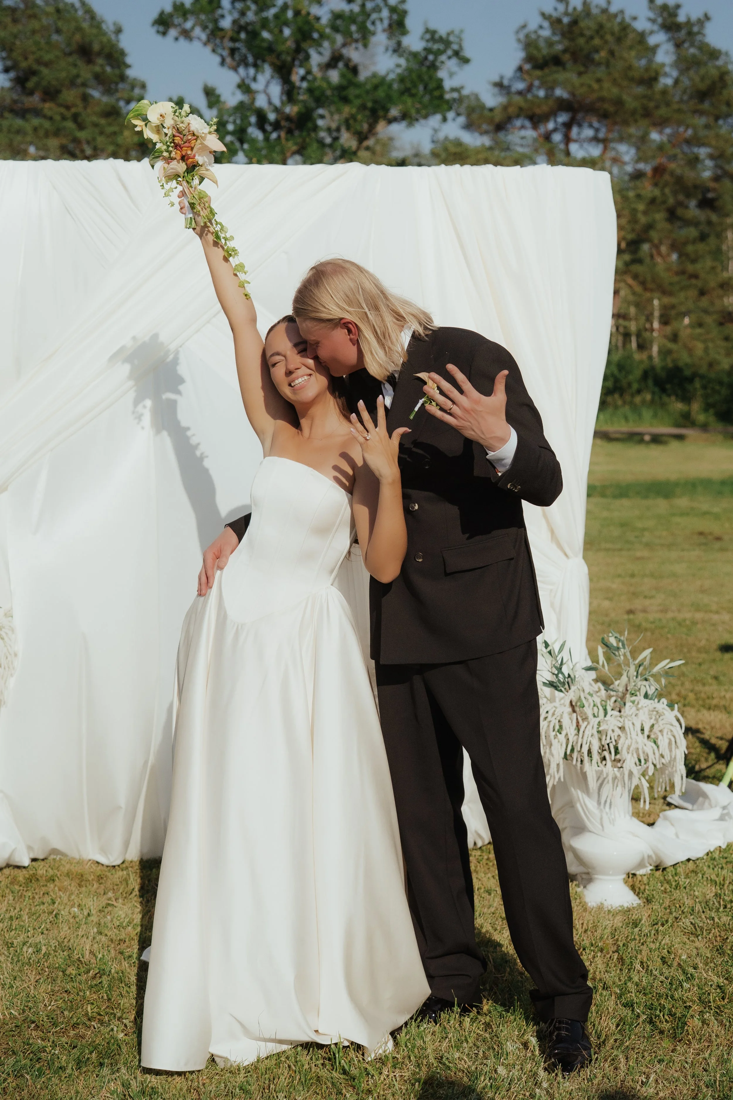 A bride and groom celebrating outdoors with a white backdrop, trees, and grass. The bride, in a white dress, holds a bouquet in the air, and the groom, in a black suit, kisses her on the cheek.