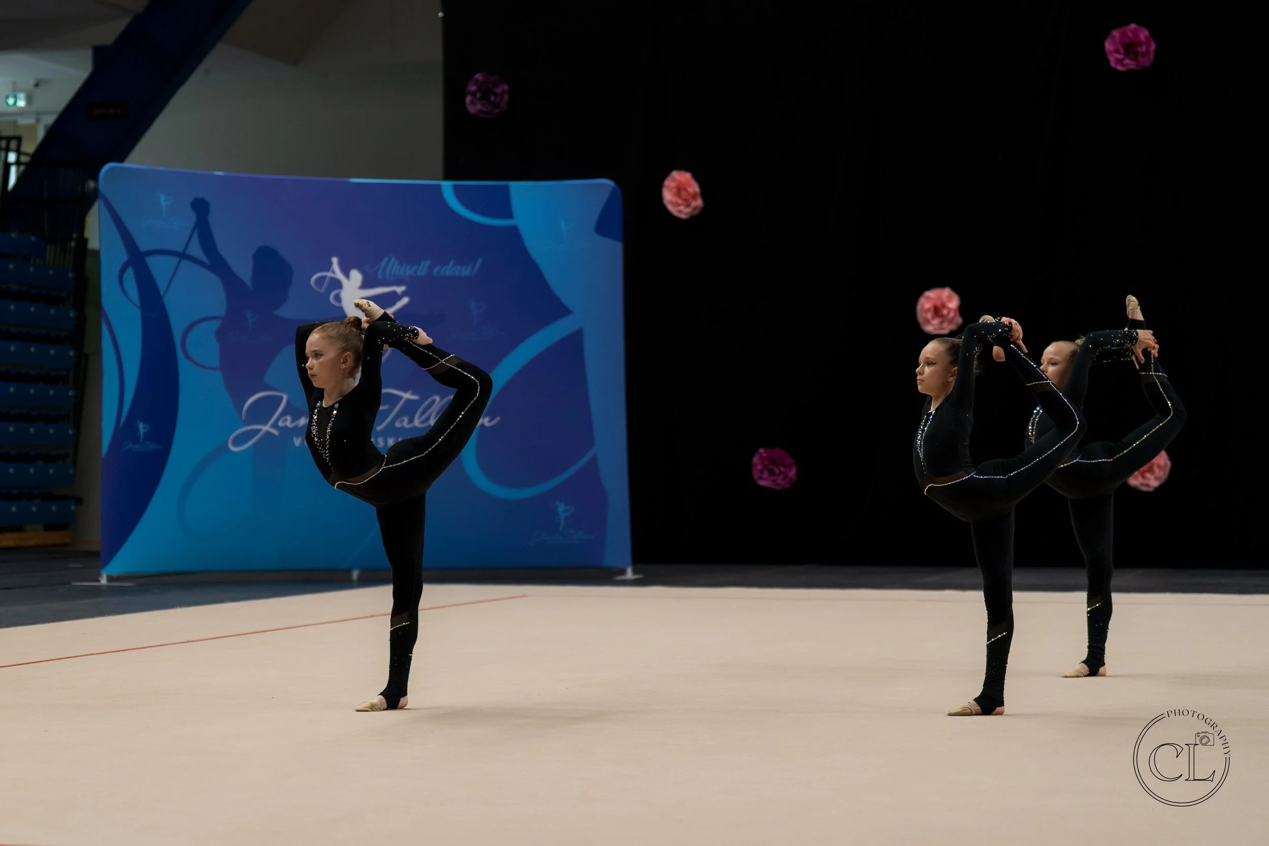 Three young female gymnasts in black leotards performing a synchronized balance pose during a gymnastics routine on a light-colored mat in an indoor sports arena.
