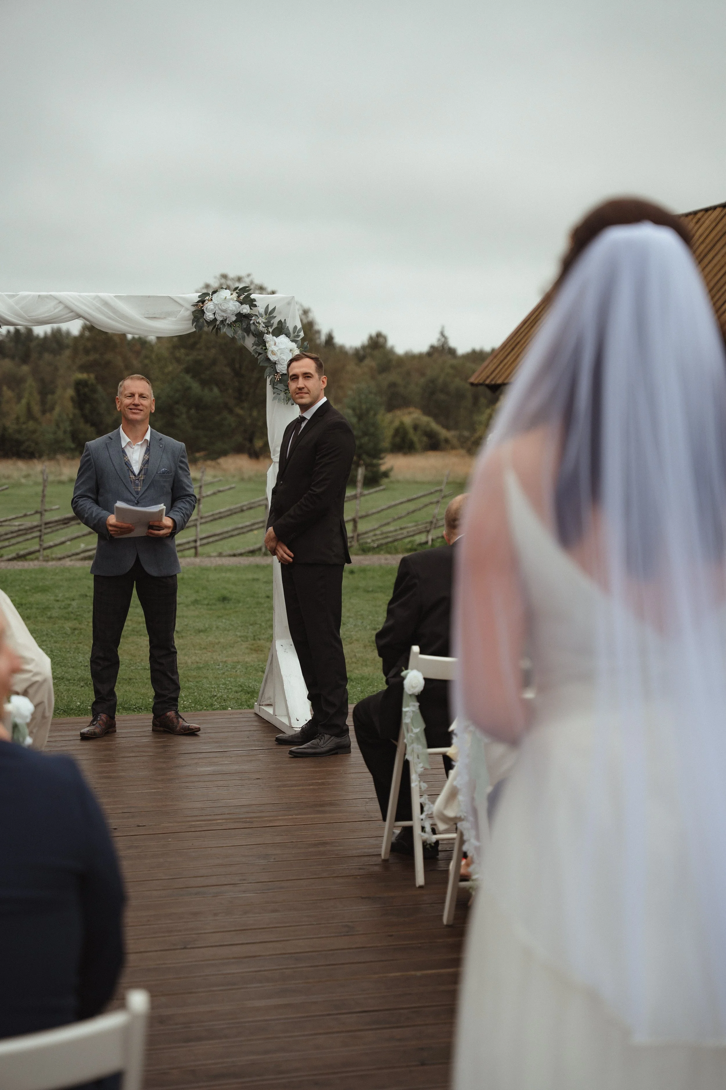 An outdoor wedding ceremony with a bride in a white dress and veil, a groom in a black suit, and an officiant in a blue blazer standing on a wooden platform, with guests seated in front and a floral wedding arch in the background.