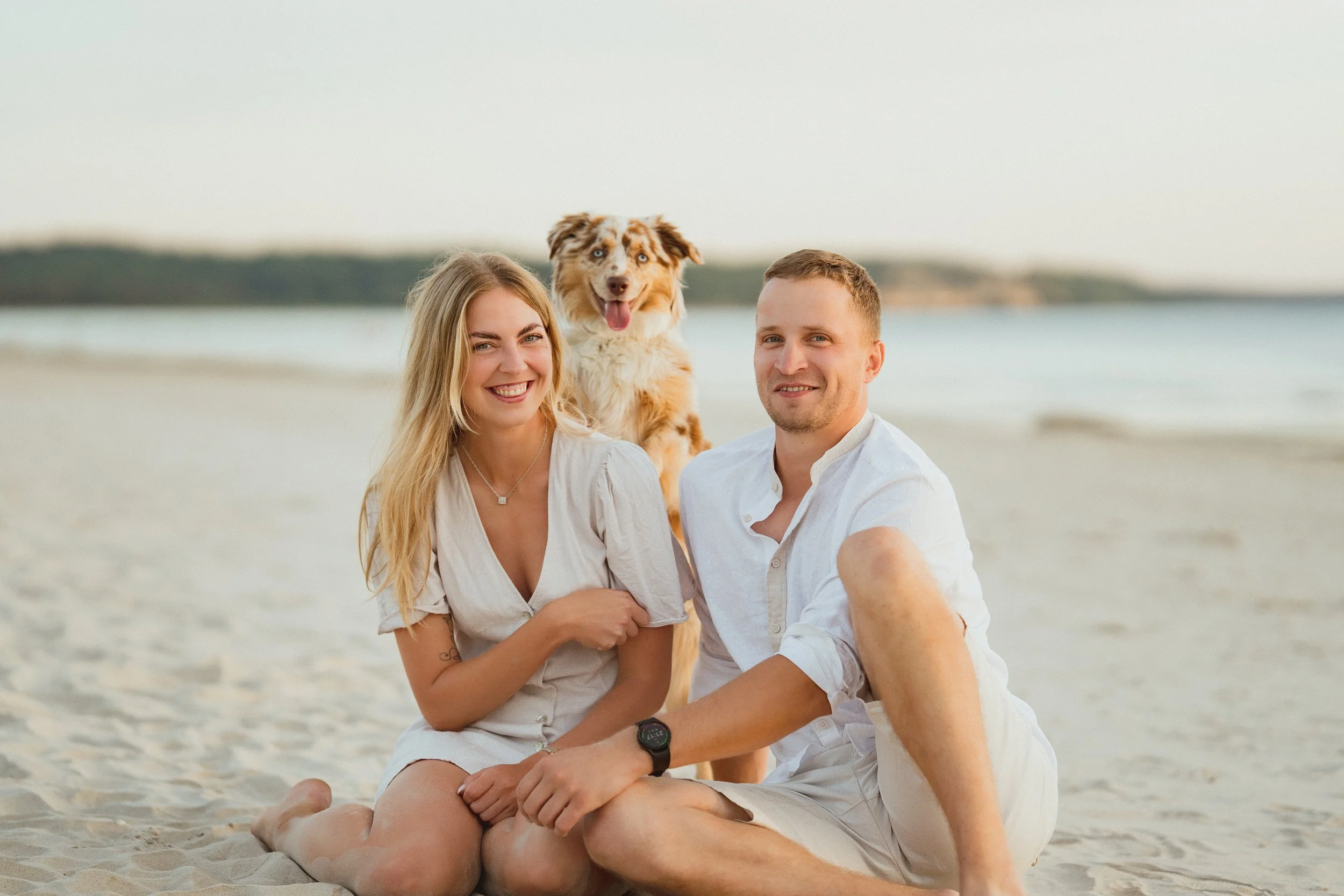 A happy couple and their Australian Shepherd dog sitting on the beach at sunset.