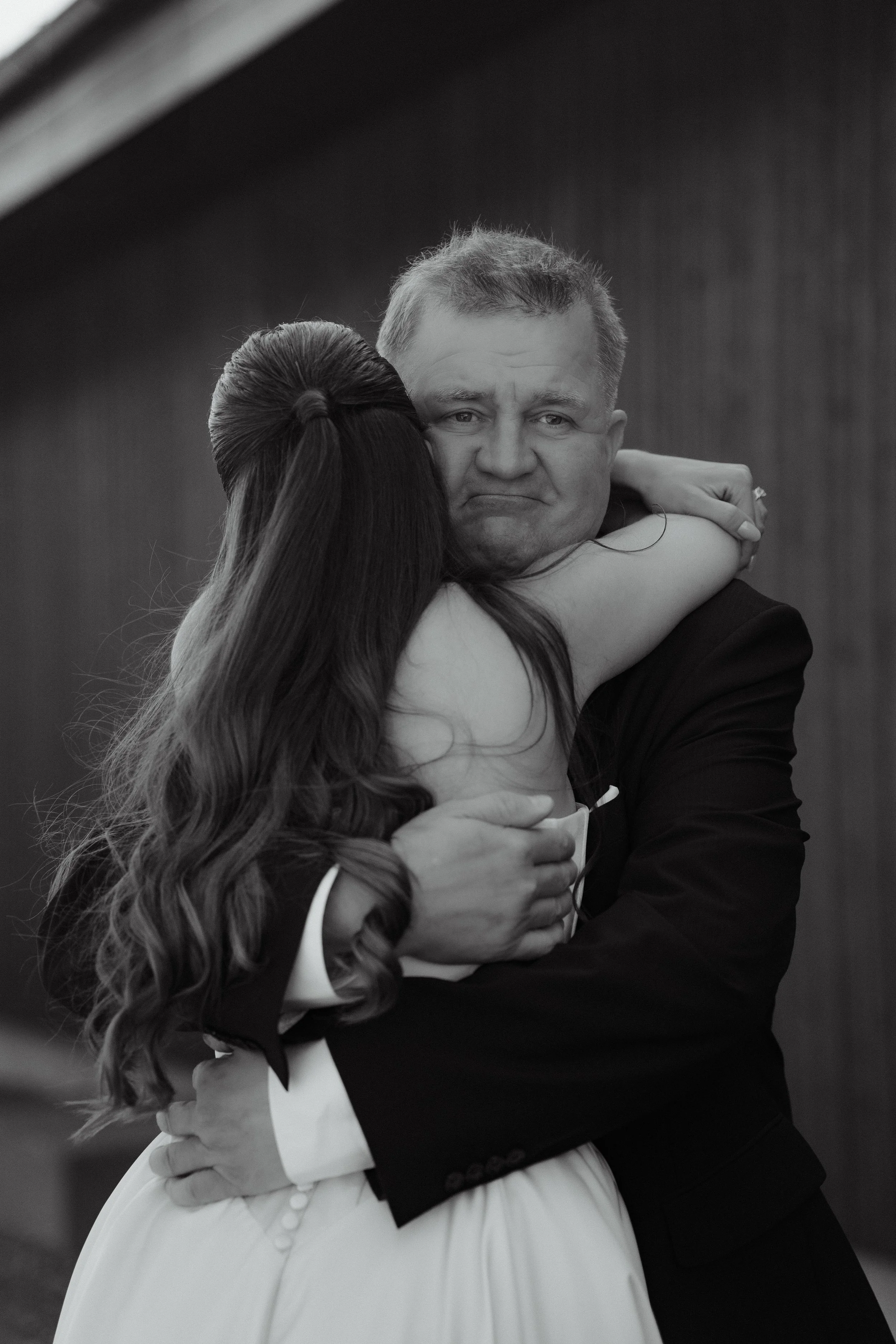 A black and white photo of a man in a tuxedo hugging a woman in a white dress, the woman is facing away and the man has a sad or emotional expression.