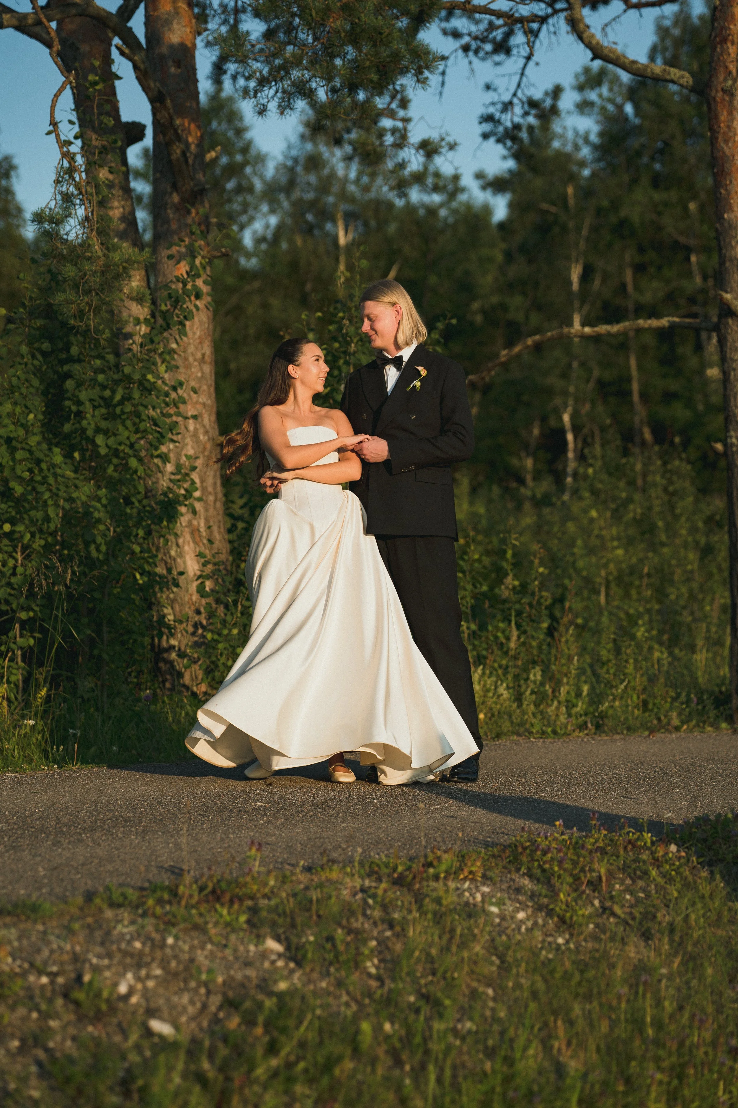 A couple in wedding attire, holding hands and smiling at each other, standing outdoors on a paved path in a wooded area during sunset.