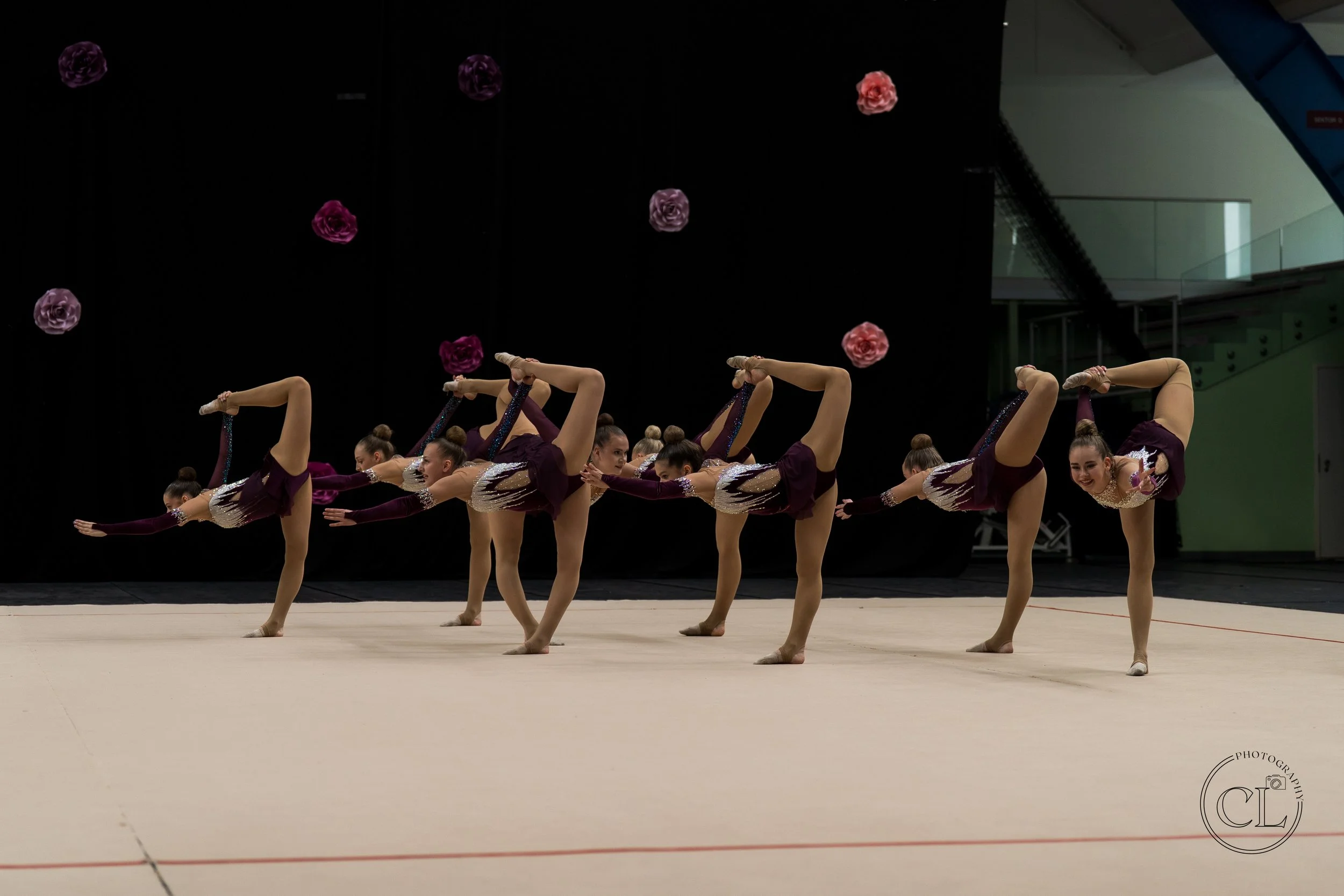 Group of young female gymnasts in purple and white costumes performing a synchronized routine on a gymnastics floor, each balancing on one leg while holding the other leg behind their head with one arm extended forward.